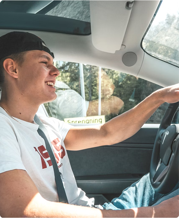 Young man with a black cap smiling and driving a car, wearing a white T-shirt and a seatbelt.