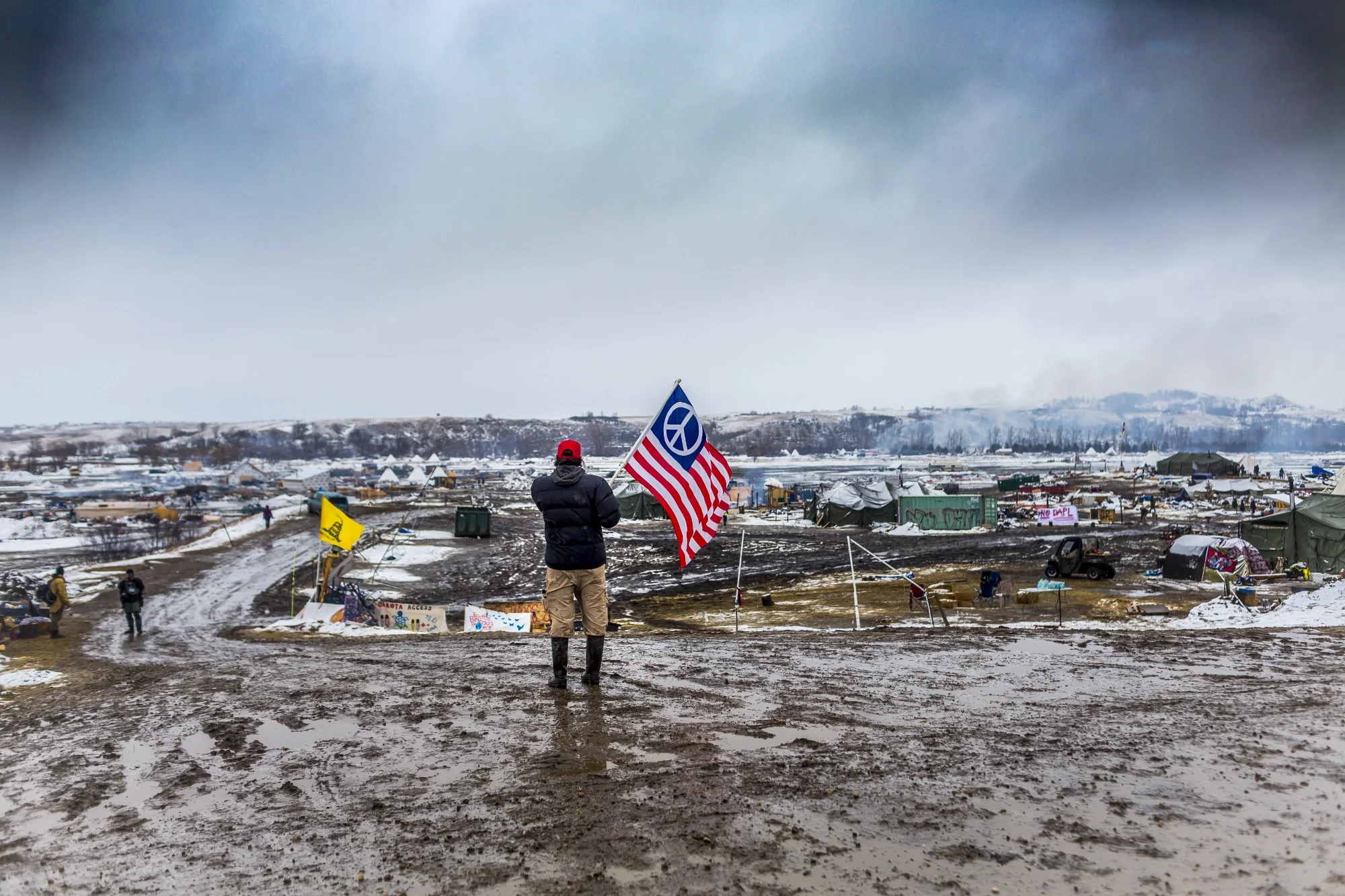 a man holds a flag while looking at the vast encampment of Standing Rock