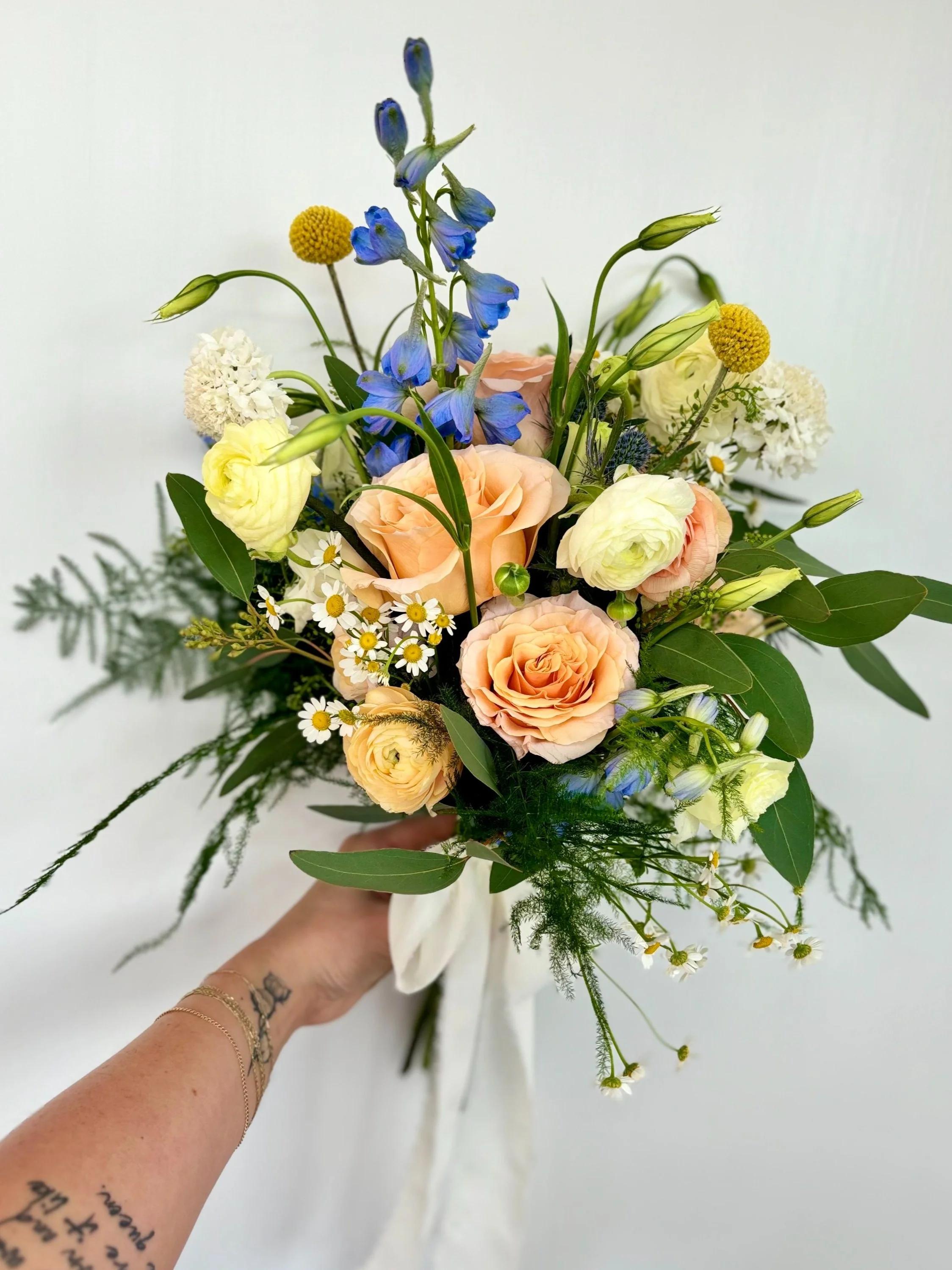Hand holding a flower bouquet with peach roses, white lisianthus, blue delphiniums, chamomile, and greenery against a white background.