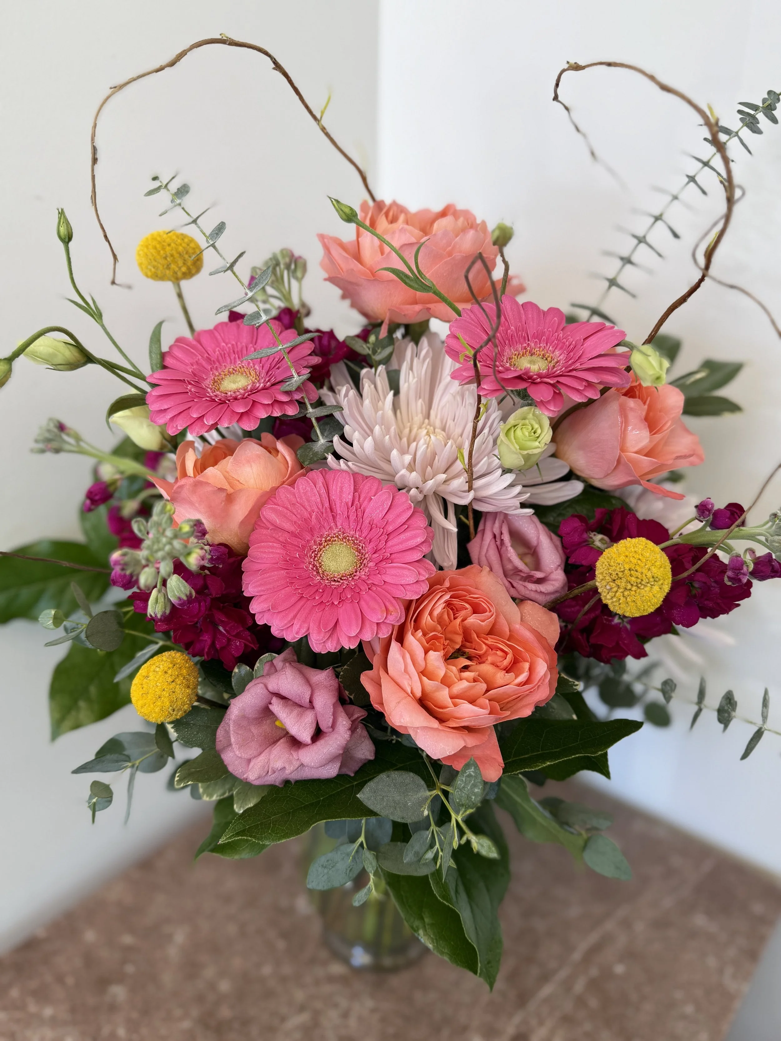 A colorful bouquet of flowers in a glass vase, featuring pink gerbera daisies, peach roses, white chrysanthemums, yellow billy balls, purple statice, and greenery, placed on a brown surface against a plain white background.