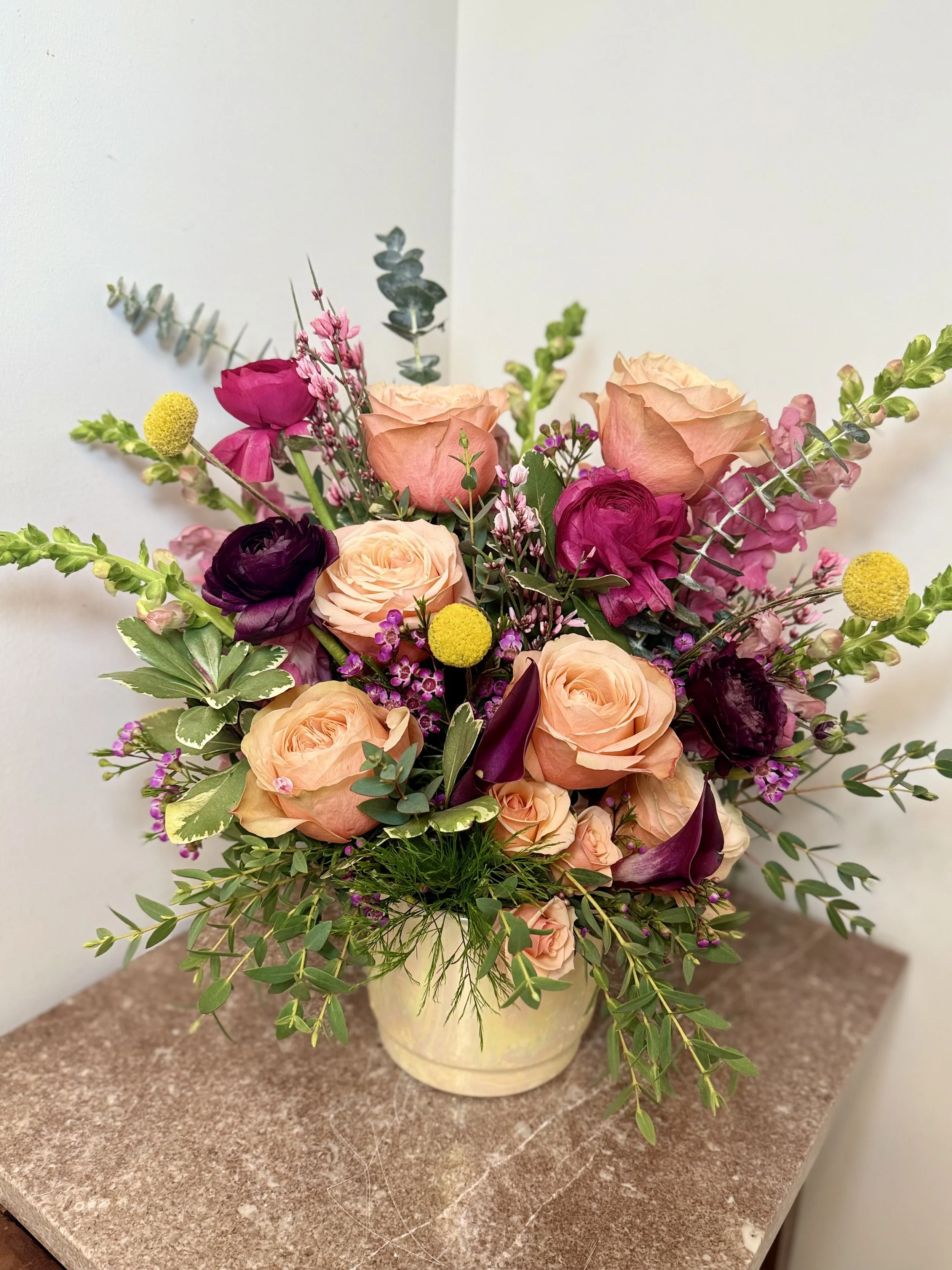 A floral arrangement with roses in peach and pink, dark purple ranunculus, yellow billy balls, pink snapdragons, and various green foliage in a cream-colored vase on a brown marble surface.
