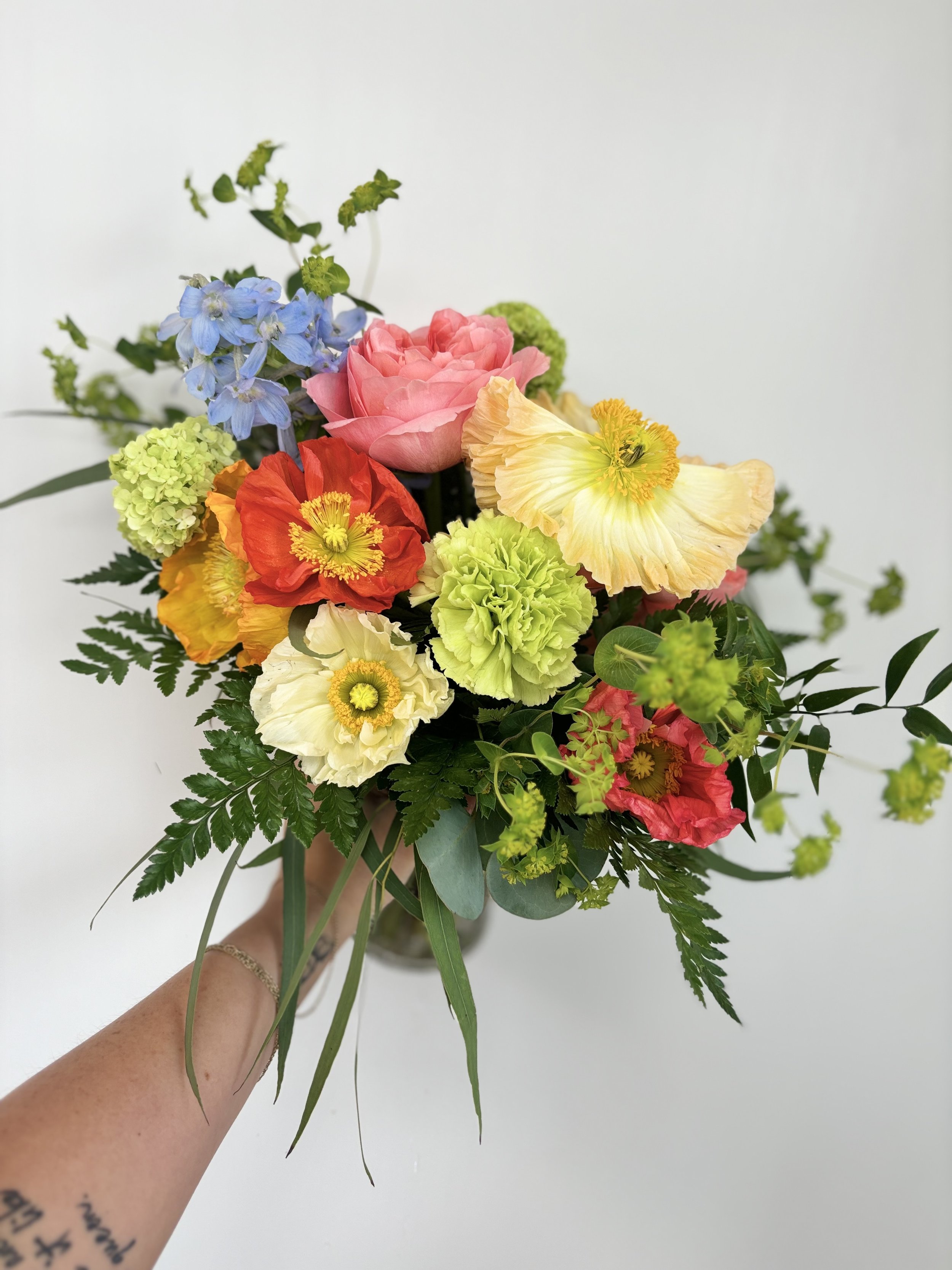 Colorful mixed flower bouquet including pink roses, blue delphiniums, yellow poppies, green carnations, and assorted greenery.