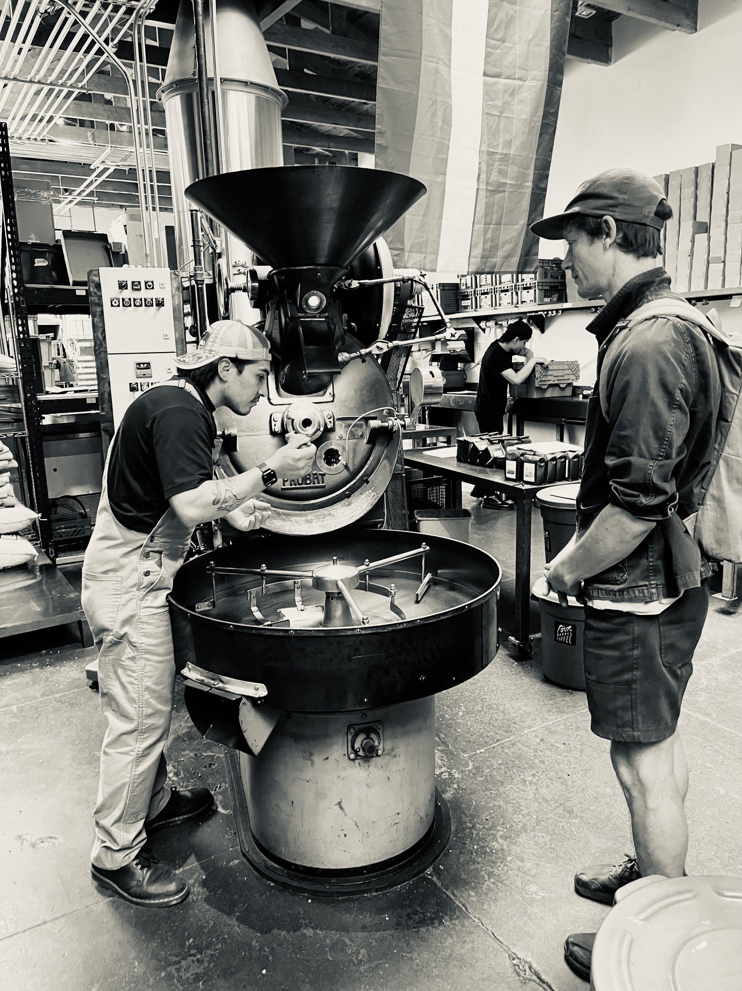 Two men working with coffee roasting equipment in a workshop or factory setting, with a woman in the background organizing items on a shelf.