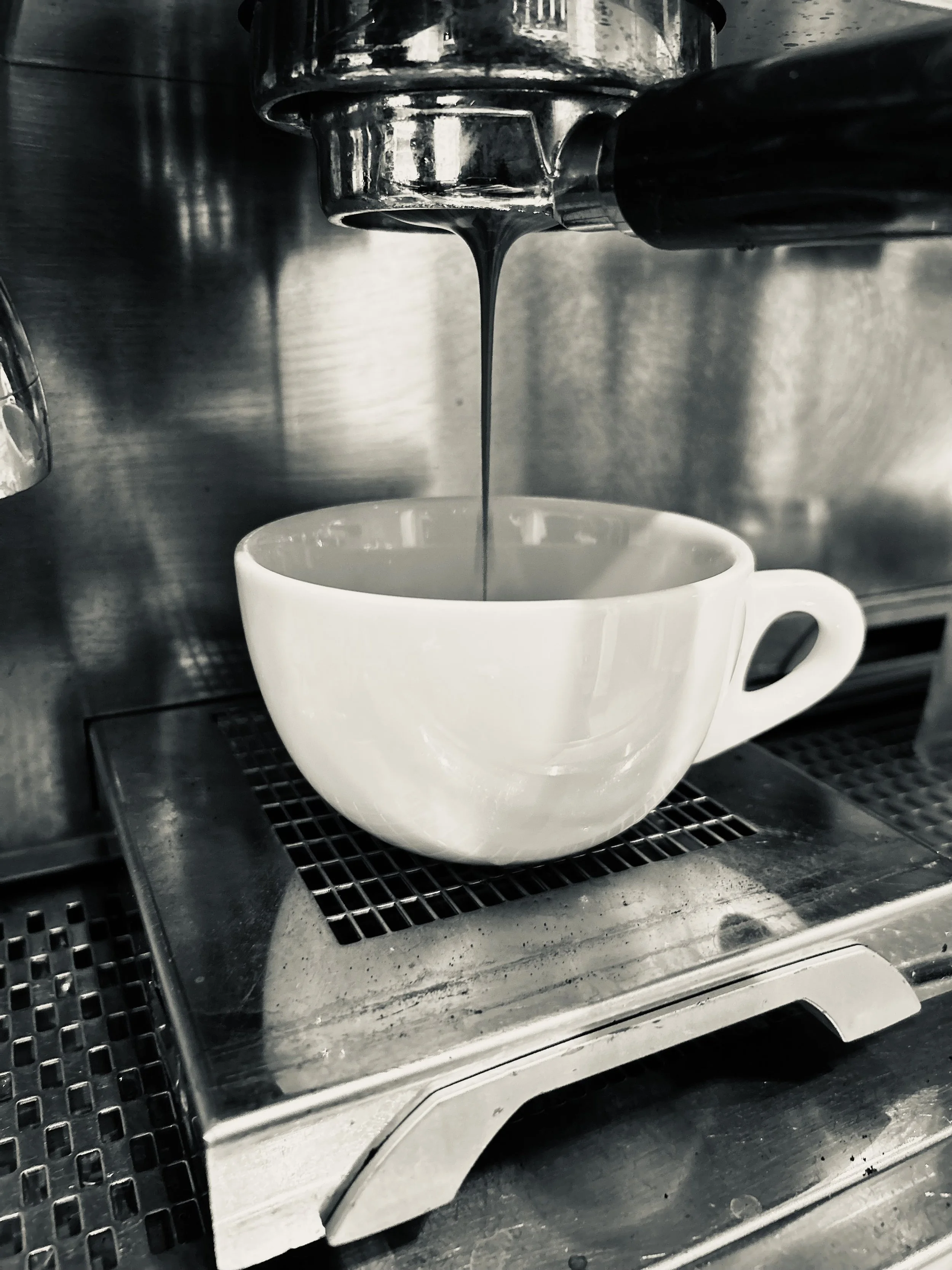Black and white photo of an espresso machine pouring coffee into a white ceramic cup on a metal drip tray.