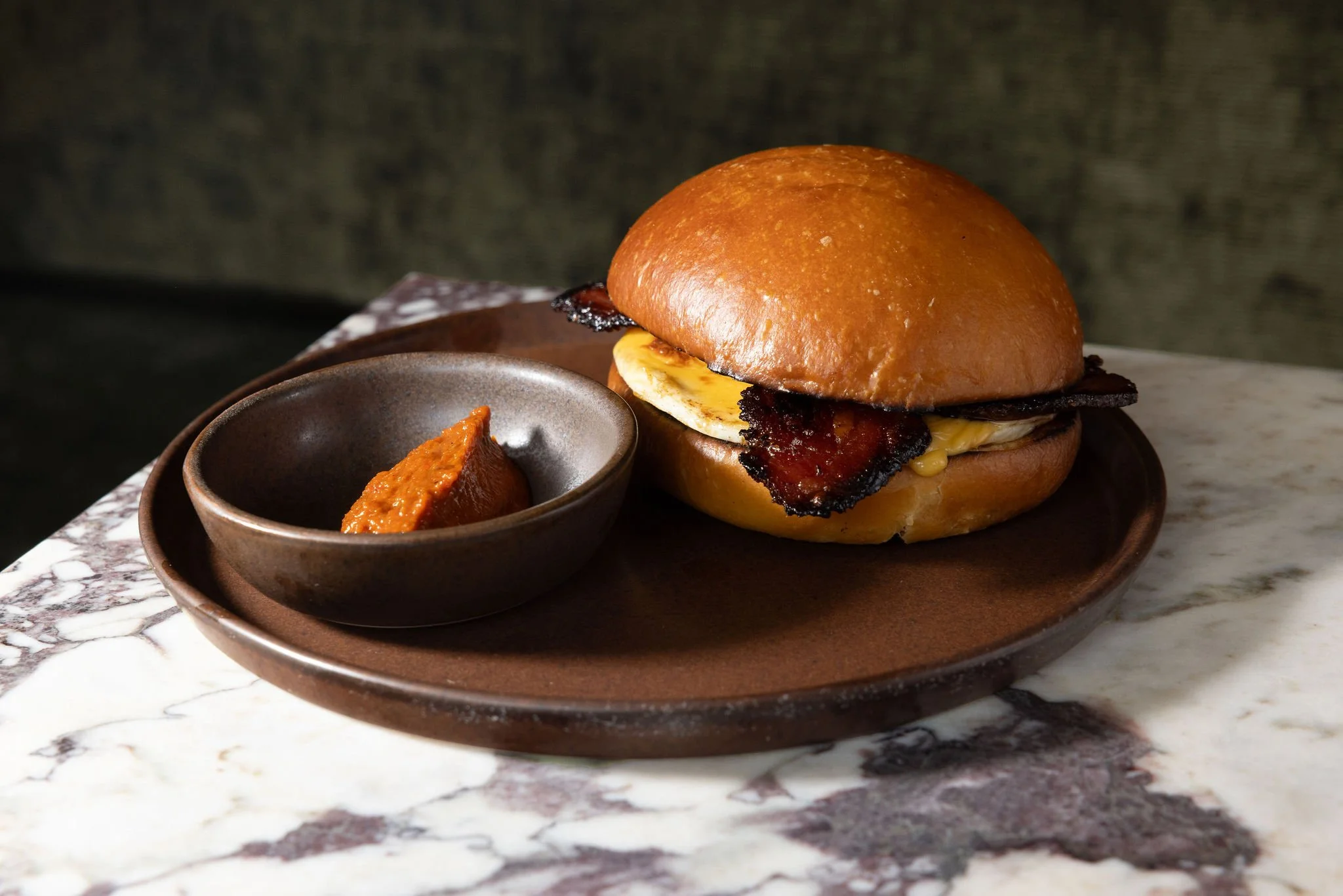 A bacon, egg, and cheese sandwich on a plate wtih a side of harissa
