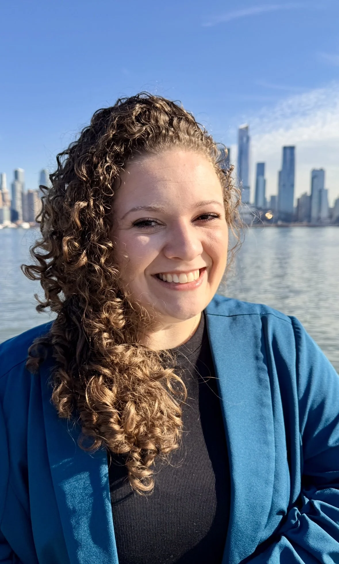 A young woman with curly hair smiling outdoors by a waterway with a city skyline in the background, wearing a blue jacket.