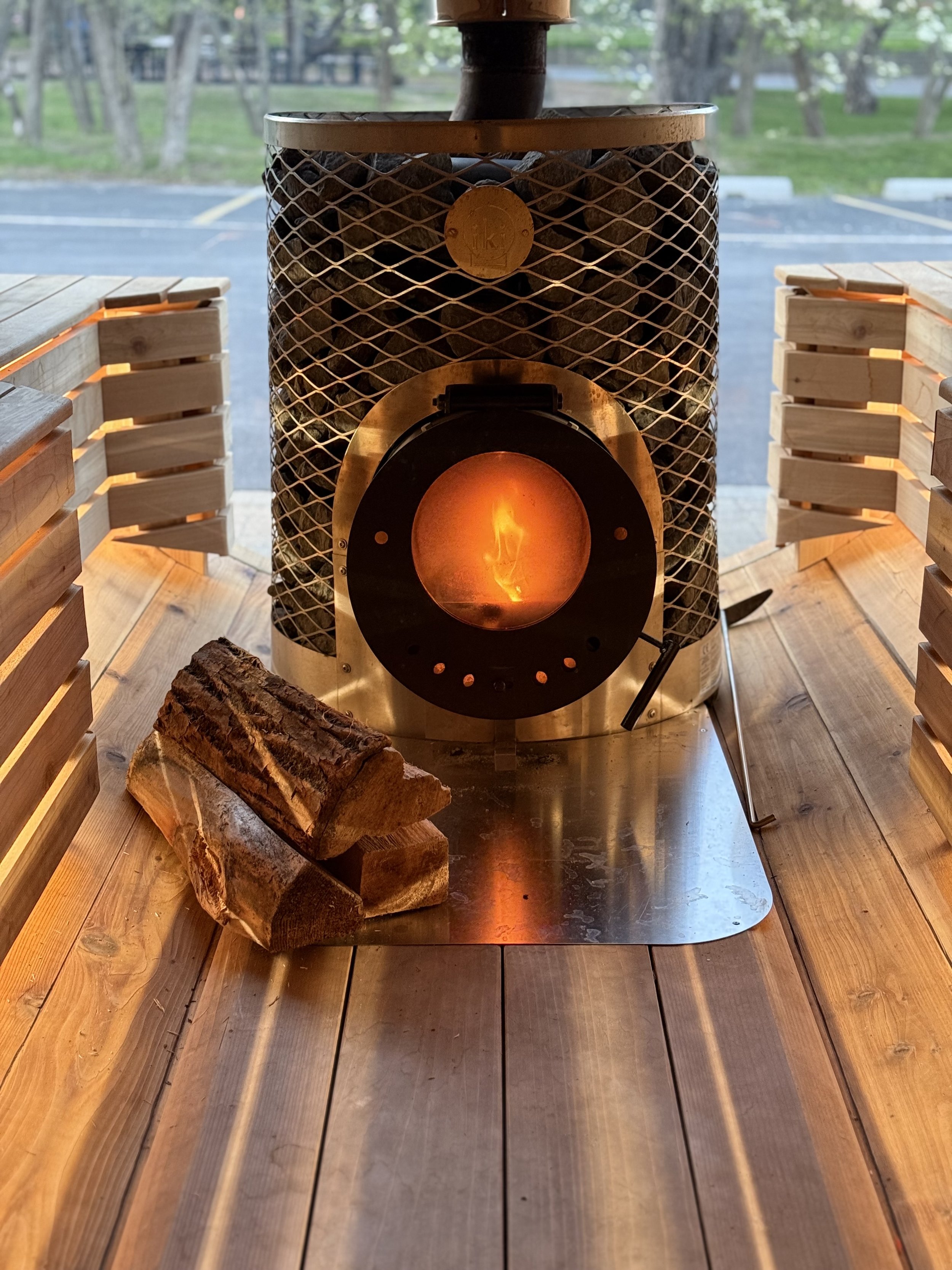 Wood-burning stove with visible flames and stacked firewood on a wooden floor.