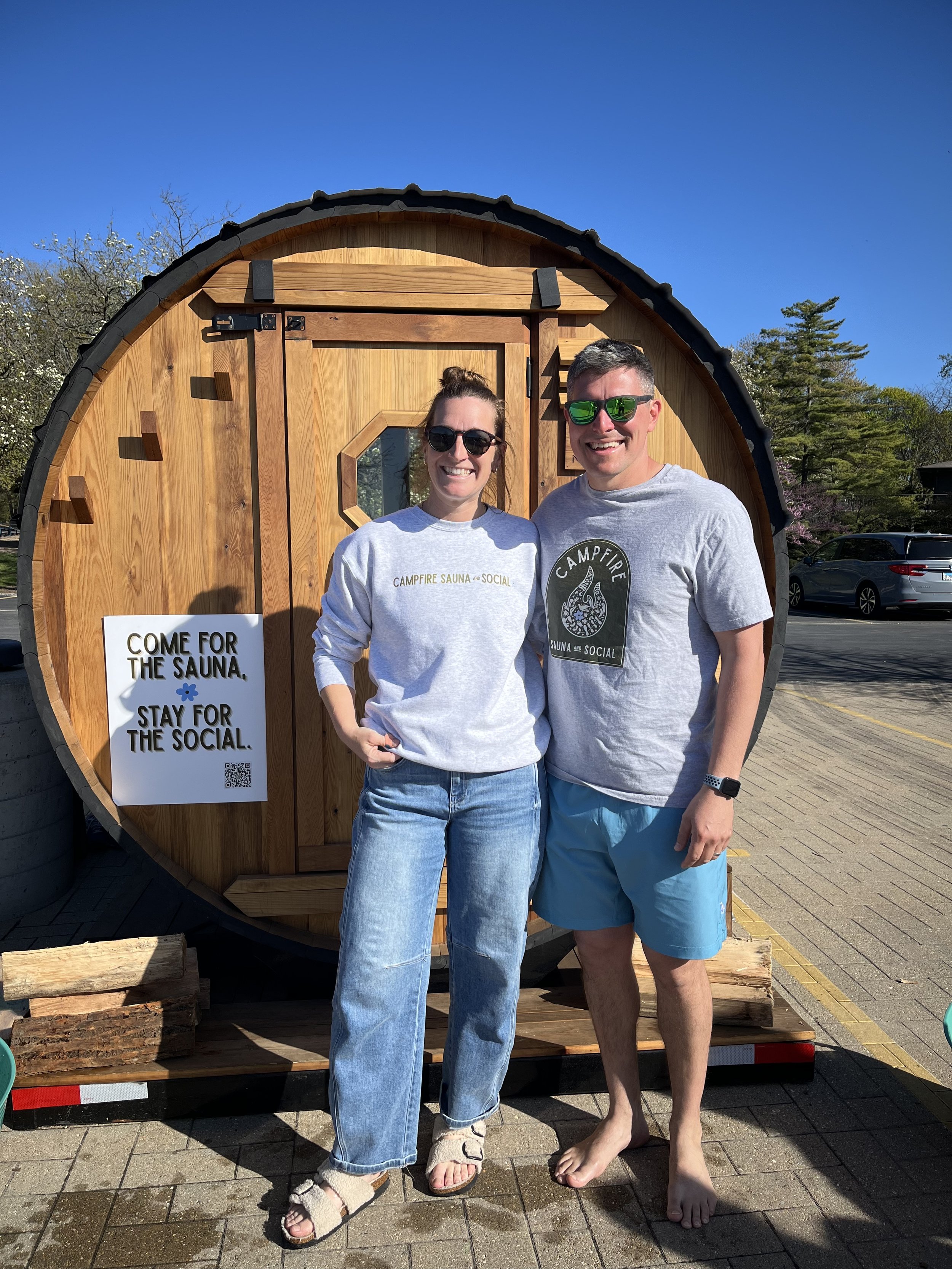 Two people standing in front of a barrel-shaped wooden sauna, smiling. They are casually dressed, with one in a sweatshirt and jeans, and the other in a t-shirt and shorts. Next to them is a sign that reads, "Come for the sauna, stay for the social."