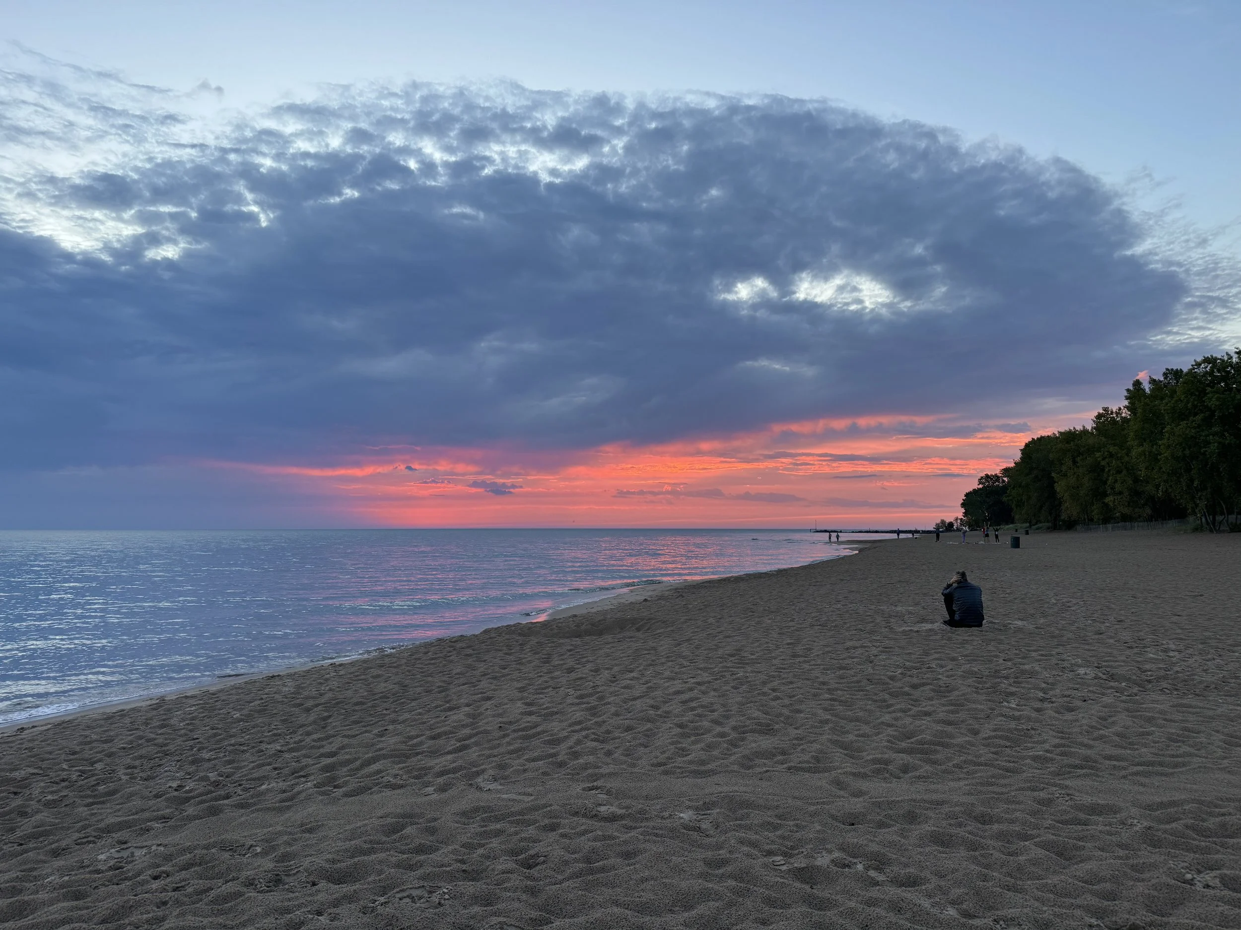 Beach at sunset with colorful clouds, calm water, and a person sitting on the sand taking a photo.