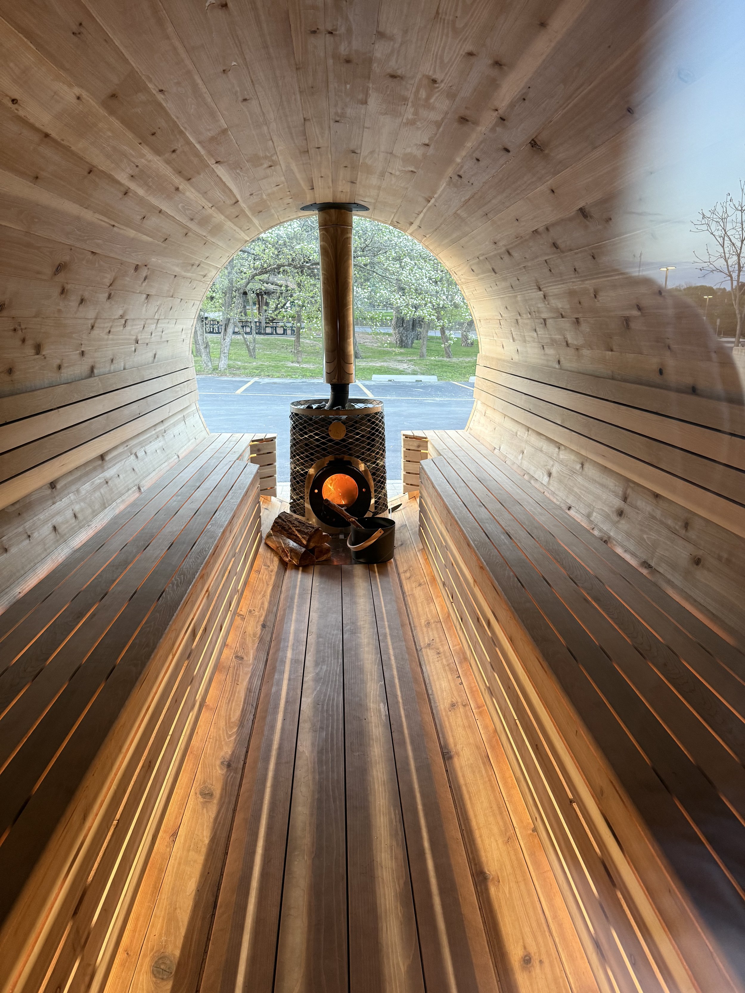 Interior of a wooden barrel sauna with benches, a chimney, and a wood stove.