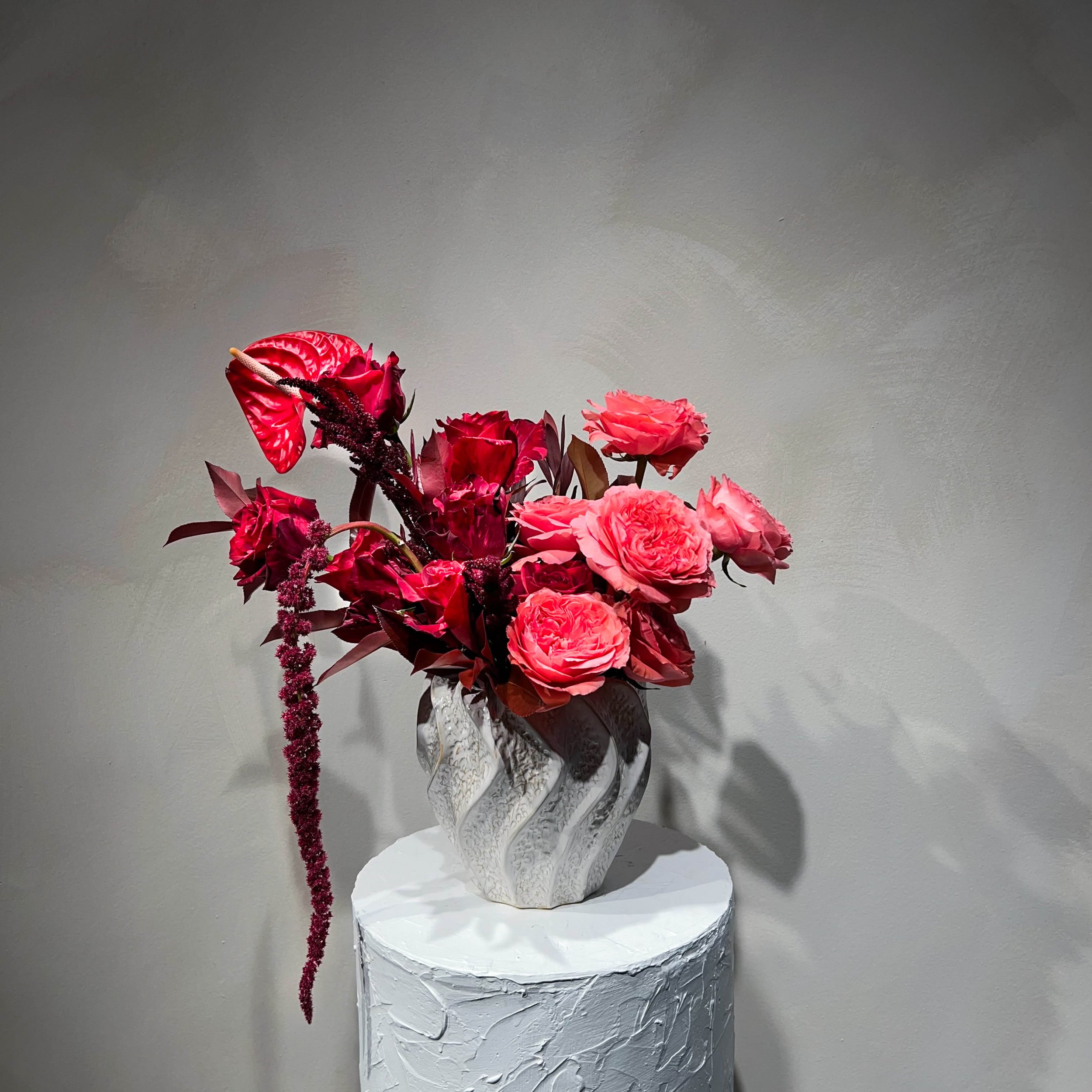 A bouquet of pink and red flowers in a textured white vase on a round white pedestal against a plain wall.