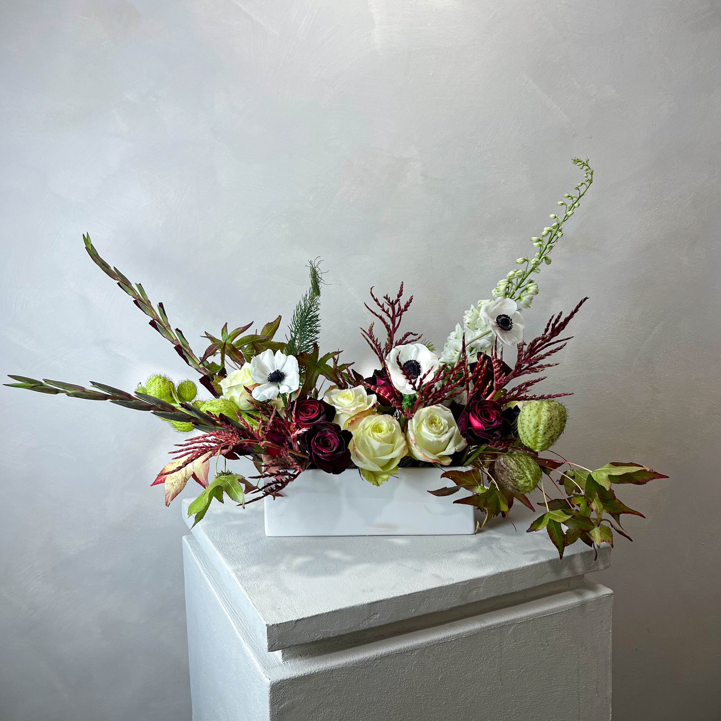 A floral arrangement with white and dark red roses, white anemones, and assorted greenery in a white rectangular vase, displayed on a white pedestal against a plain gray wall.