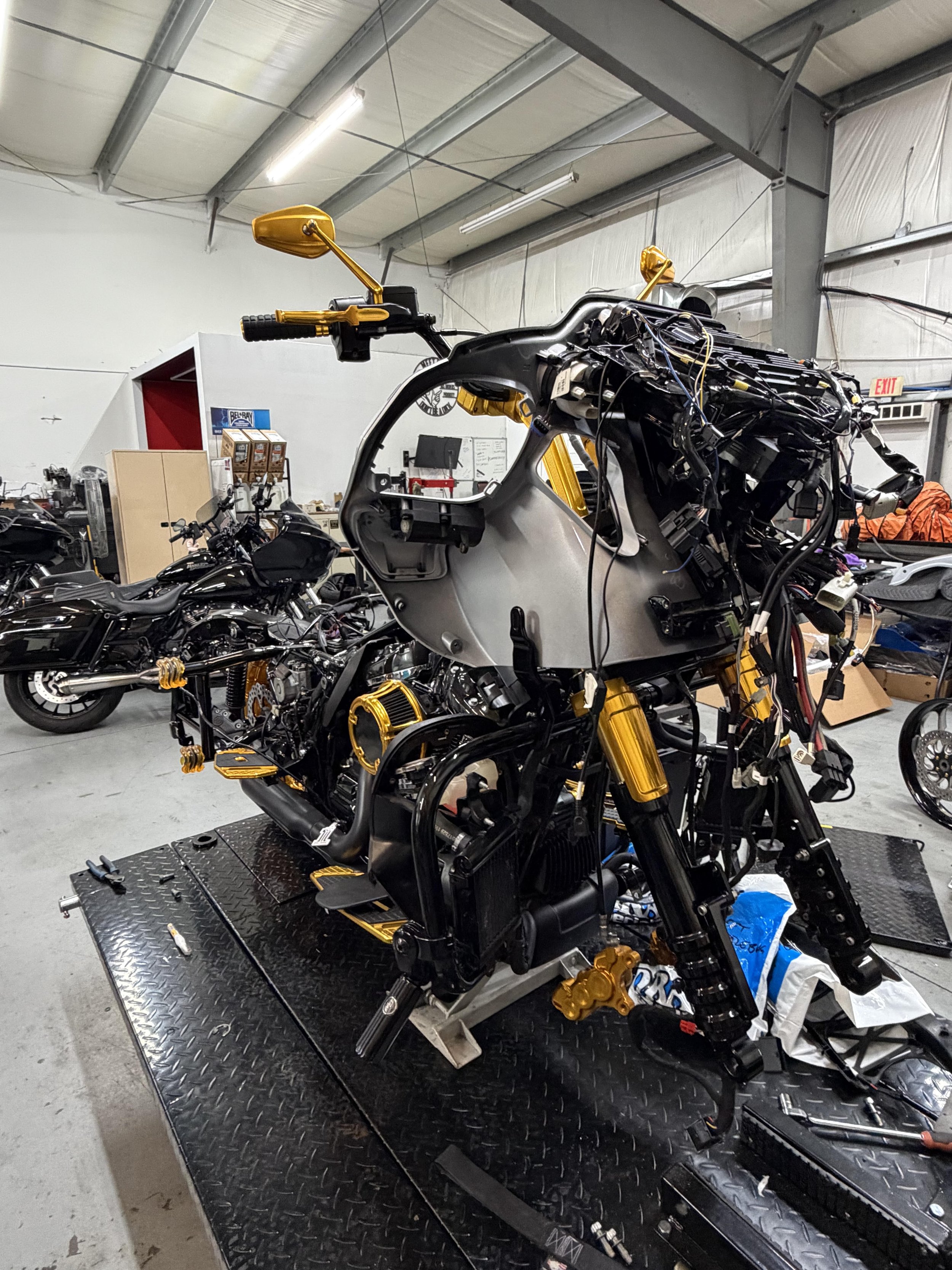 Disassembled motorcycle with parts and wiring exposed on a workbench in a workshop with motorcycles in the background.