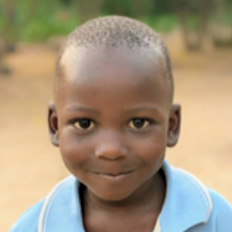 Close-up of a young boy with a short haircut, wearing a light blue shirt, smiling outdoors.