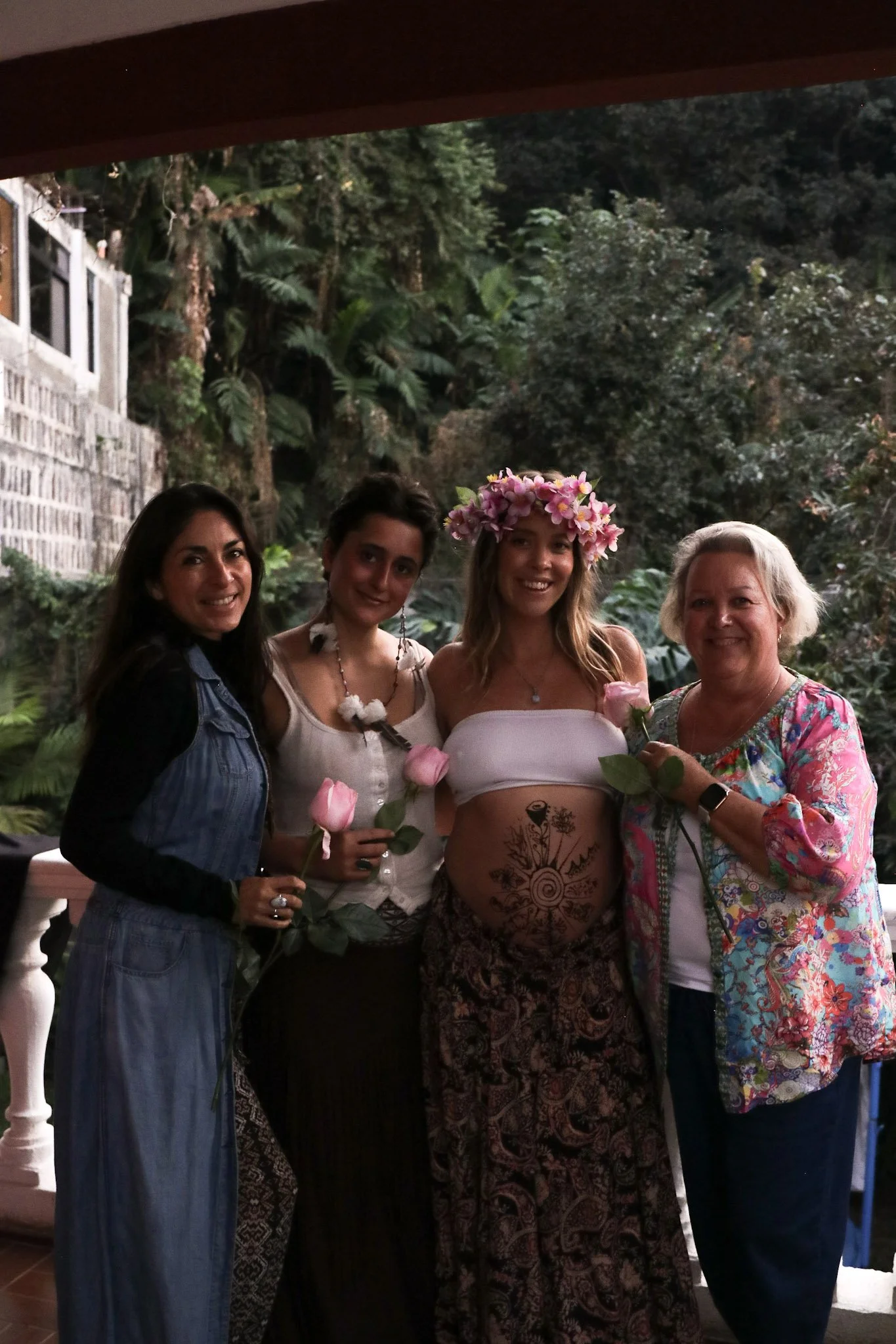 Four women standing together outdoors, one woman with a floral crown, holding pink roses, and another with tattoos on her abdomen, smiling at the camera.