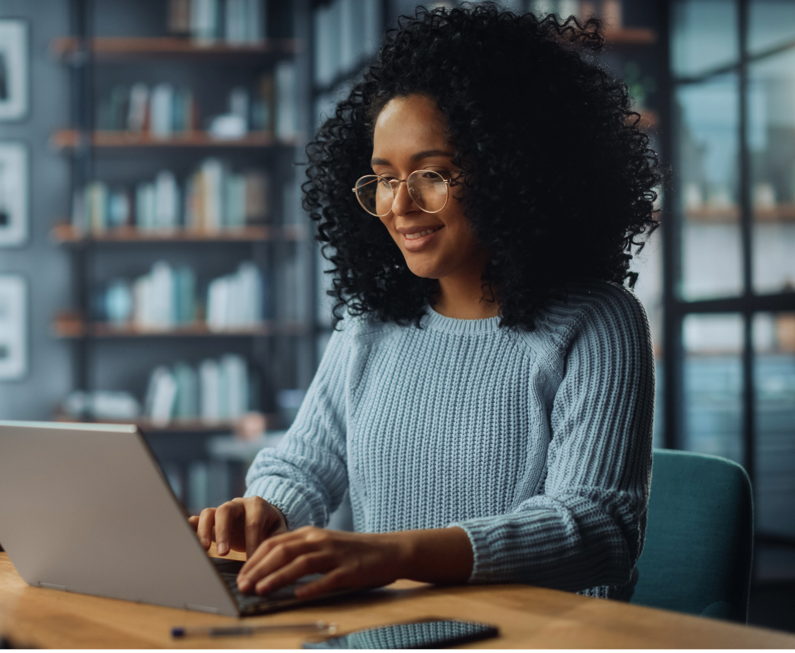 A woman with curly black hair and glasses working on a laptop in a modern office environment.