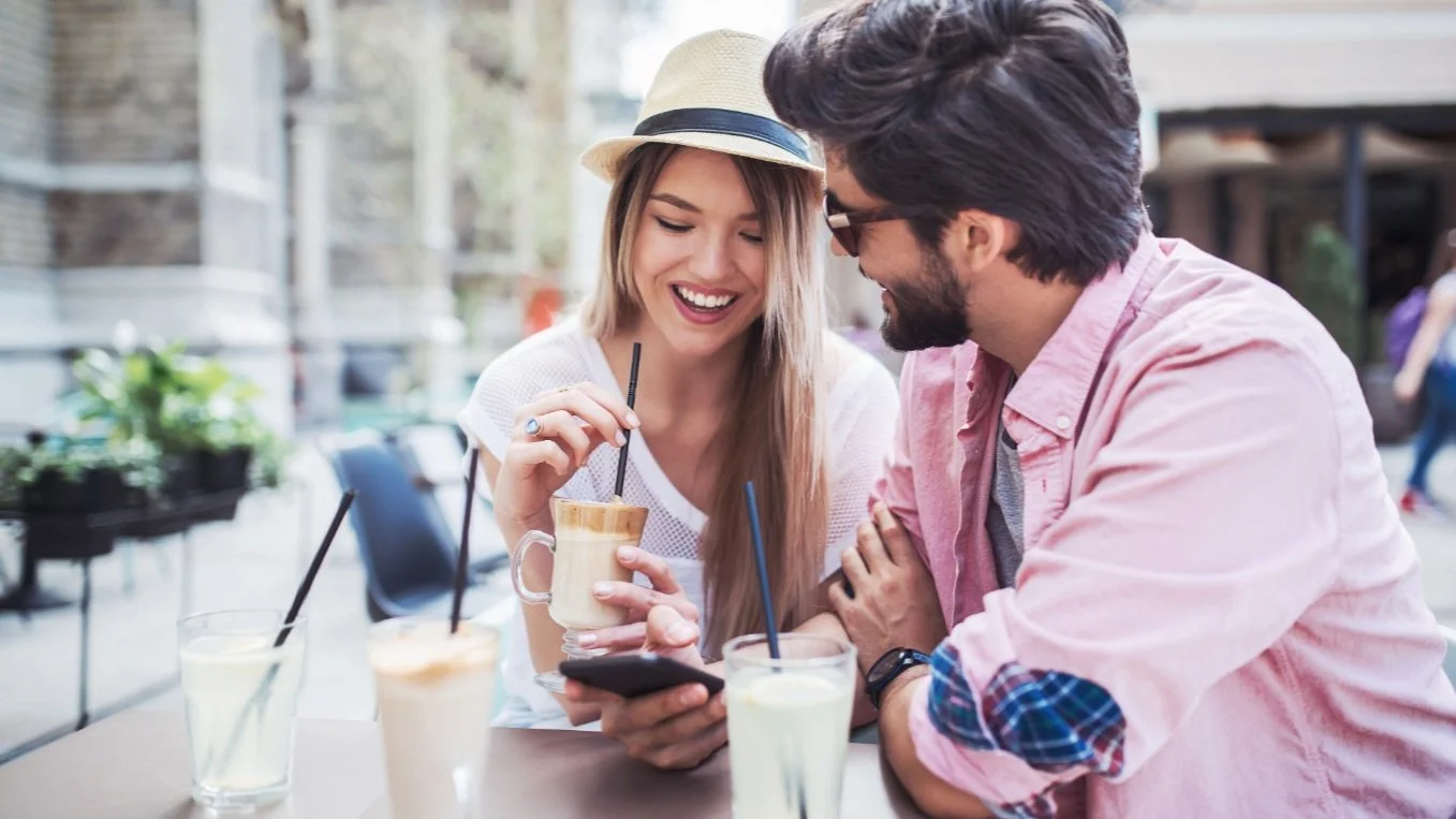 A young woman and man sit at a table outdoors, smiling and looking at a smartphone together, with several drinks in front of them.