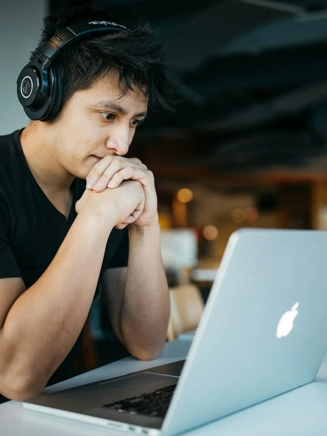 Young man with black hair wearing black headphones, sitting at a table with a silver MacBook, looking thoughtful with hands clasped in front of his mouth in a coffee shop.