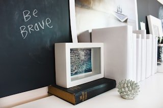 Decorative desk with framed photo, black and white books, and a small geometric sculpture, with a chalkboard saying 'Be Brave' in the background.