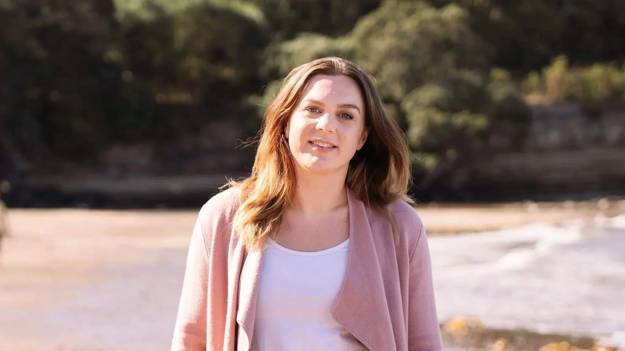 A woman with long light brown hair and blue eyes standing outdoors on a sunny day, wearing a white top and pink blazer, with trees in the background.