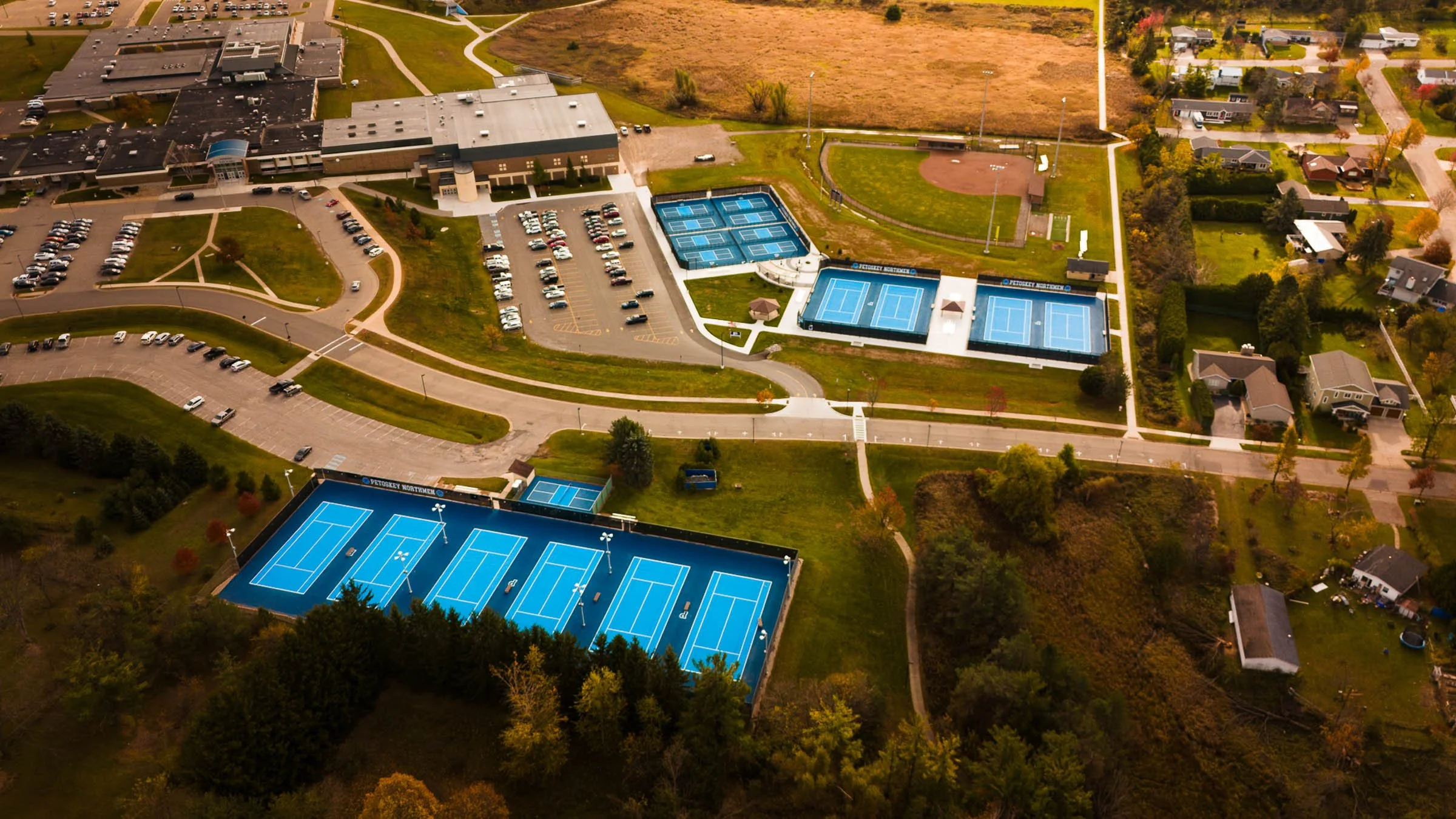 Aerial view of a recreational area in Petoskey Michigan with tennis courts, parking lots, a building, and nearby residential houses and greenery.