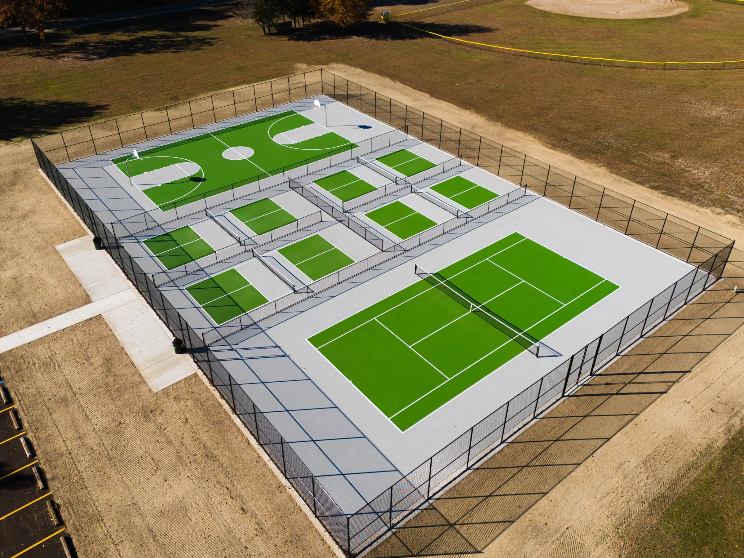 Aerial view of a multi sport facility in West Michigan featuring tennis, pickleball, and basketball courts with bright green surfaces and white lines, enclosed by a black fence, located in a park with surrounding dirt and grass areas.