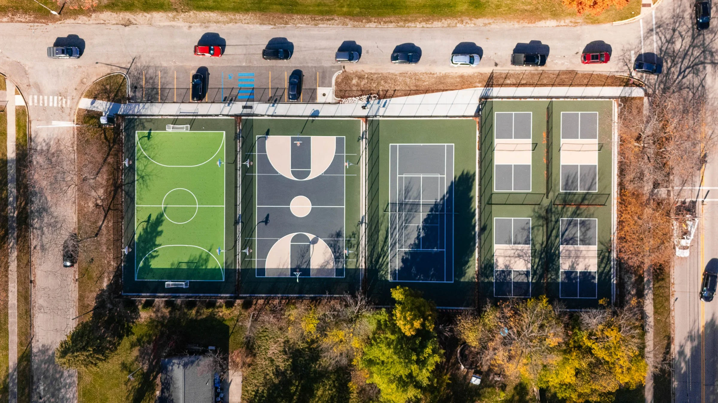 Overhead view of a sports complex in Fennville Michigan with multiple courts, including Pickleball, basketball, futsal, and tennis courts, surrounded by trees and parked cars on the street.