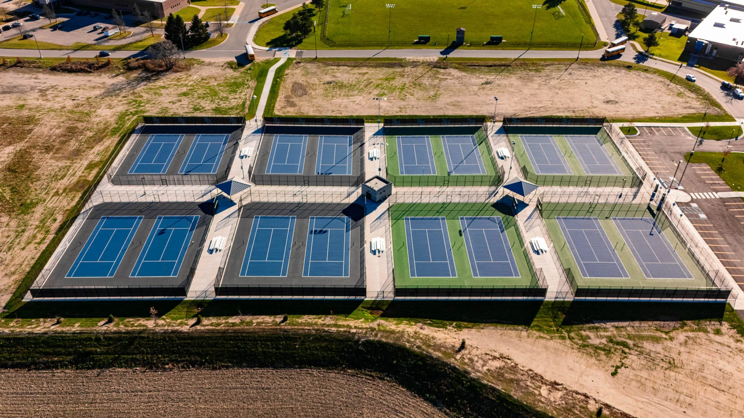 Aerial view of multiple tennis courts, some with blue surfaces and others with green surfaces, enclosed by fencing, with surrounding paved walkways and park amenities near parking lot and roads.
