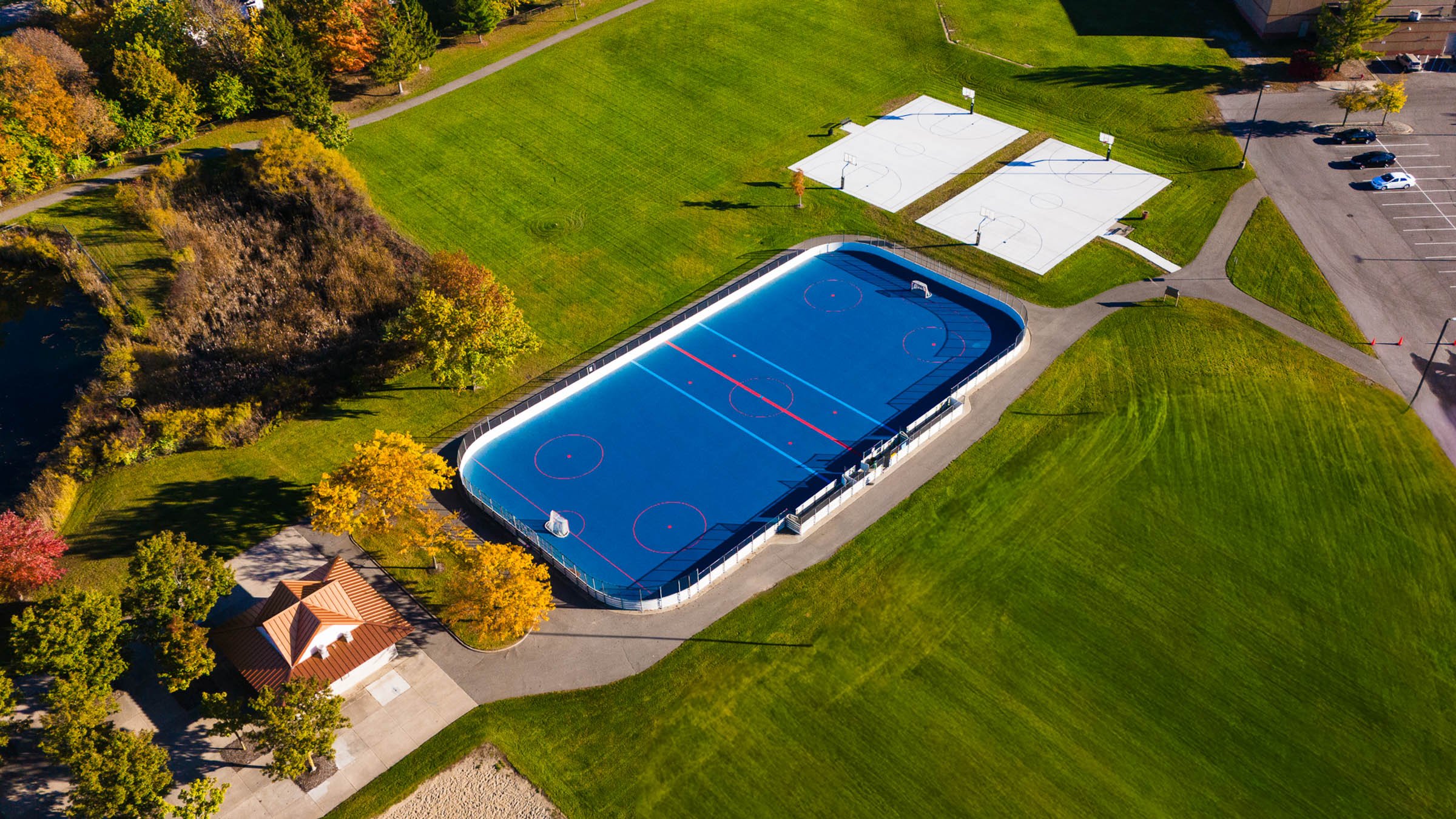 Aerial view of a sports park featuring a large ice rink, two basketball courts with nets, a parking lot with several cars, a small building, grassy areas, trees with autumn foliage, and walking paths.