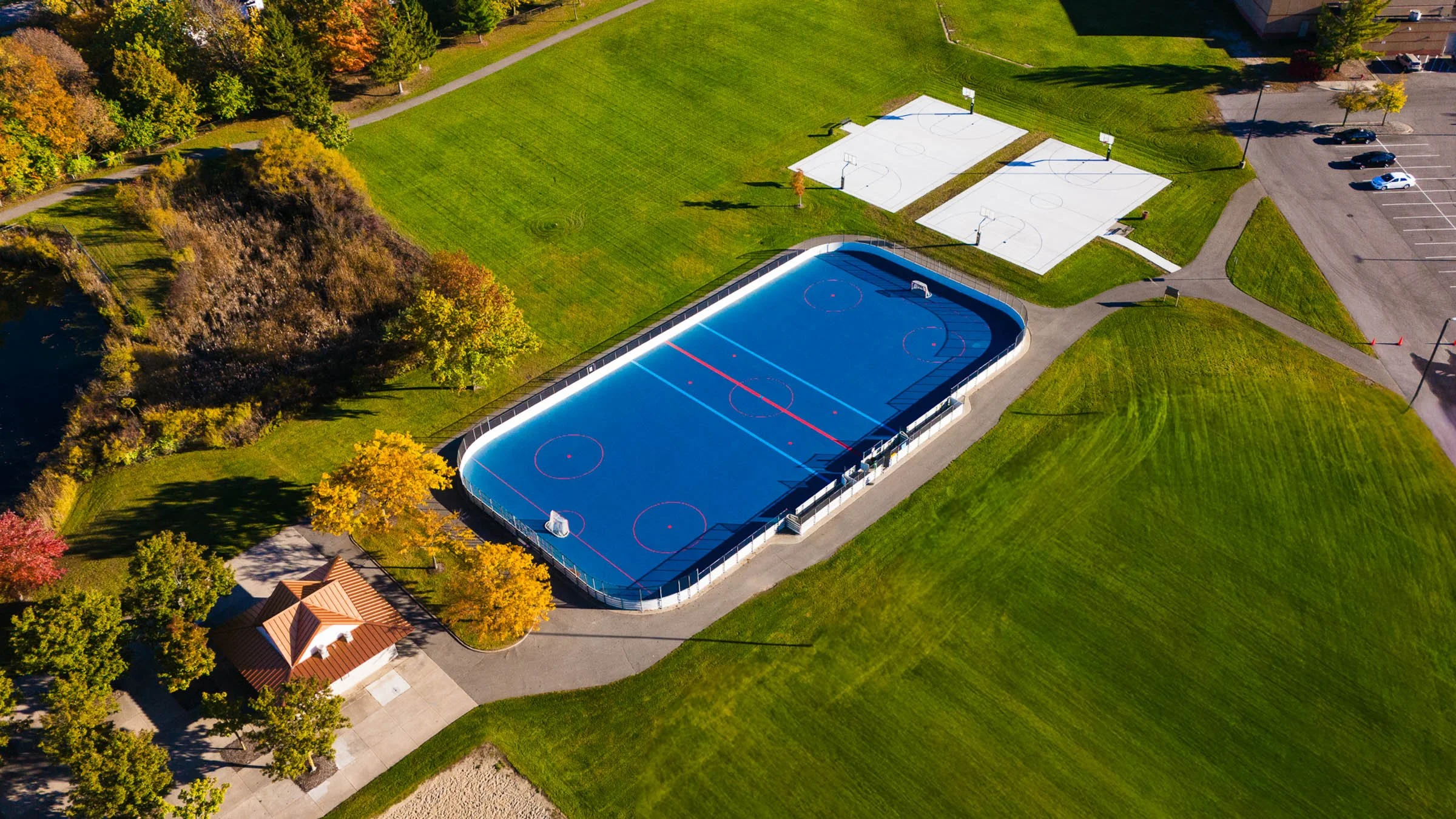 Aerial view of a sports park featuring a large ice rink, two basketball courts with nets, a parking lot with several cars, a small building, grassy areas, trees with autumn foliage, and walking paths.