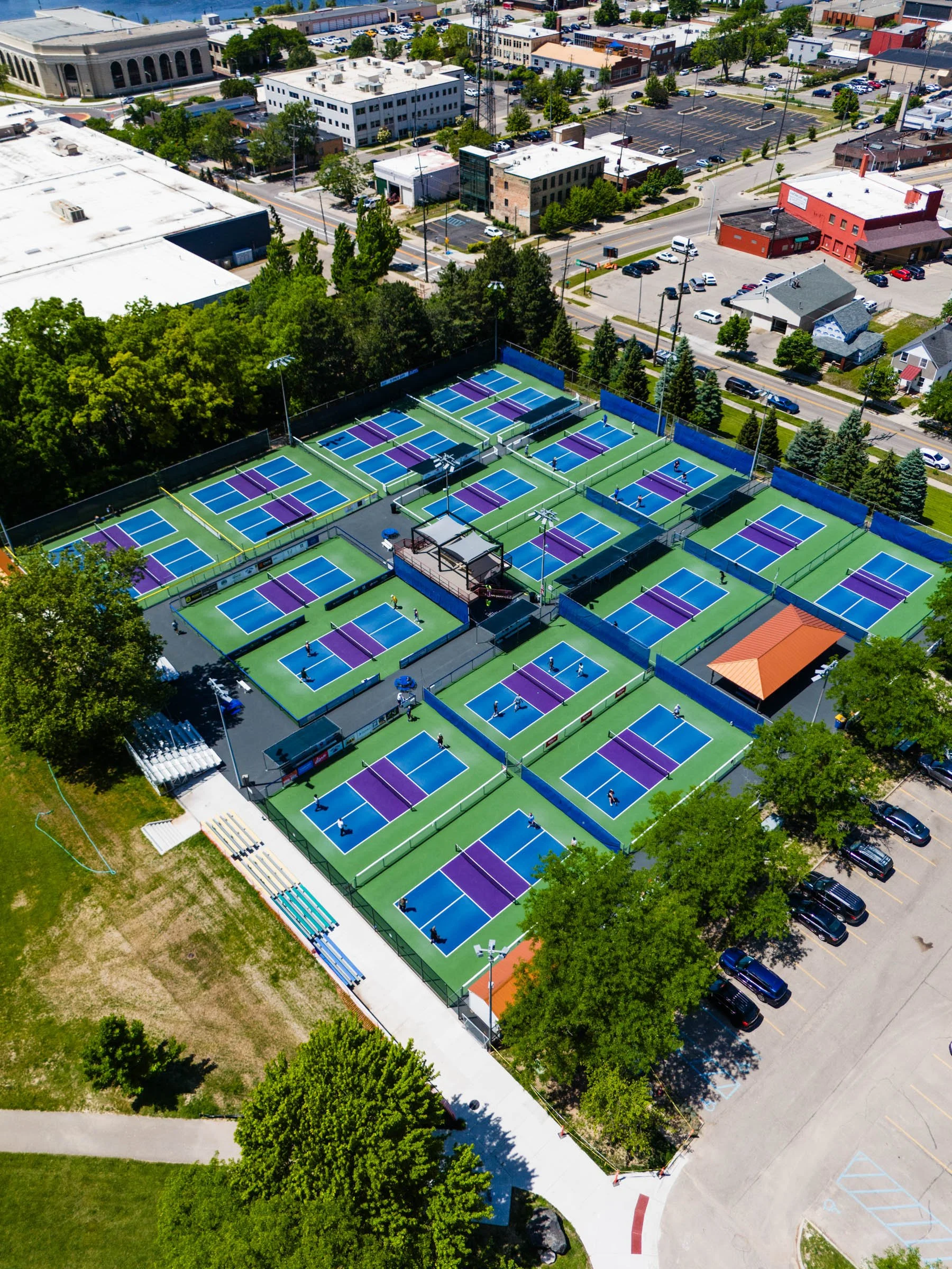 An aerial view of Belknap Park Pickleball Courts, Home of the Beer City Open, pickleball facility with multiple courts, some being used, surrounded by trees, parking lots, and urban buildings.