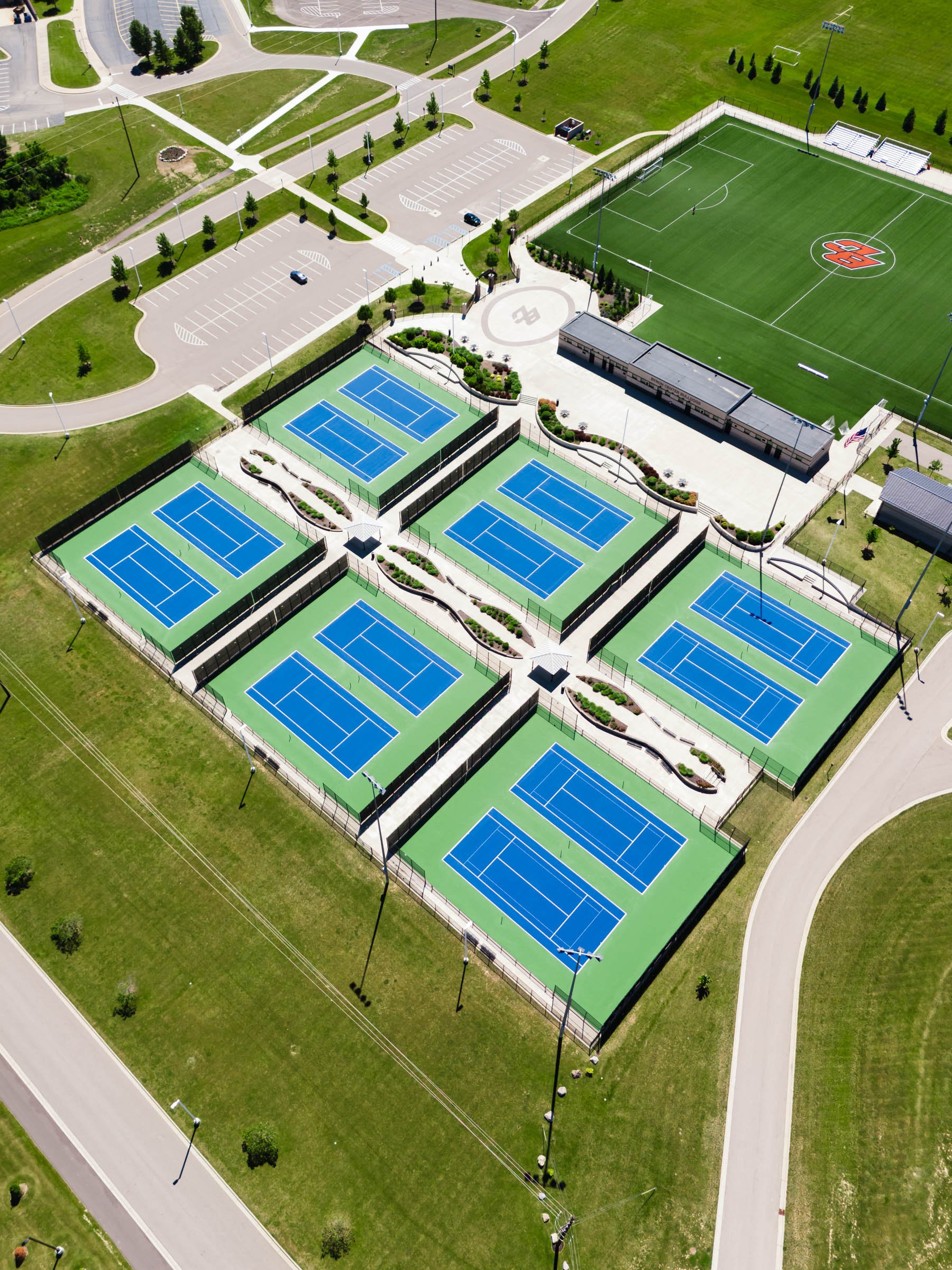 An aerial view of a sports complex, in Byron Center Michgian, with multiple tennis courts, a soccer field, parking lots, and surrounding pathways and greenery.