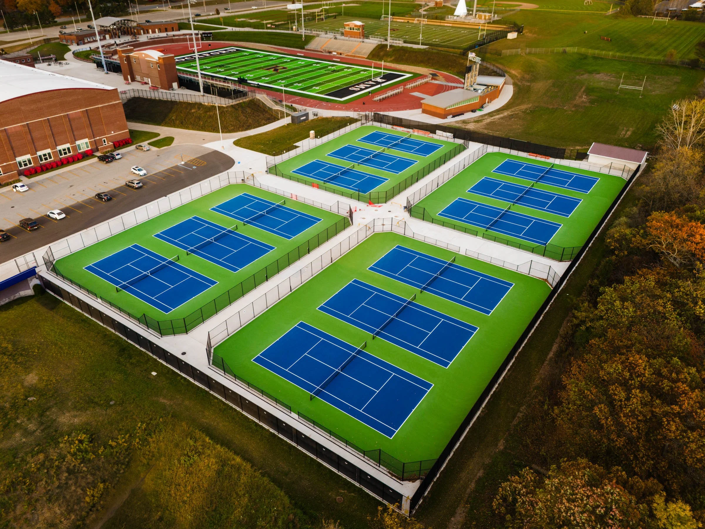 Aerial view of tennis courts with blue surfaces and surrounding green turf, located near a parking lot and sports field.