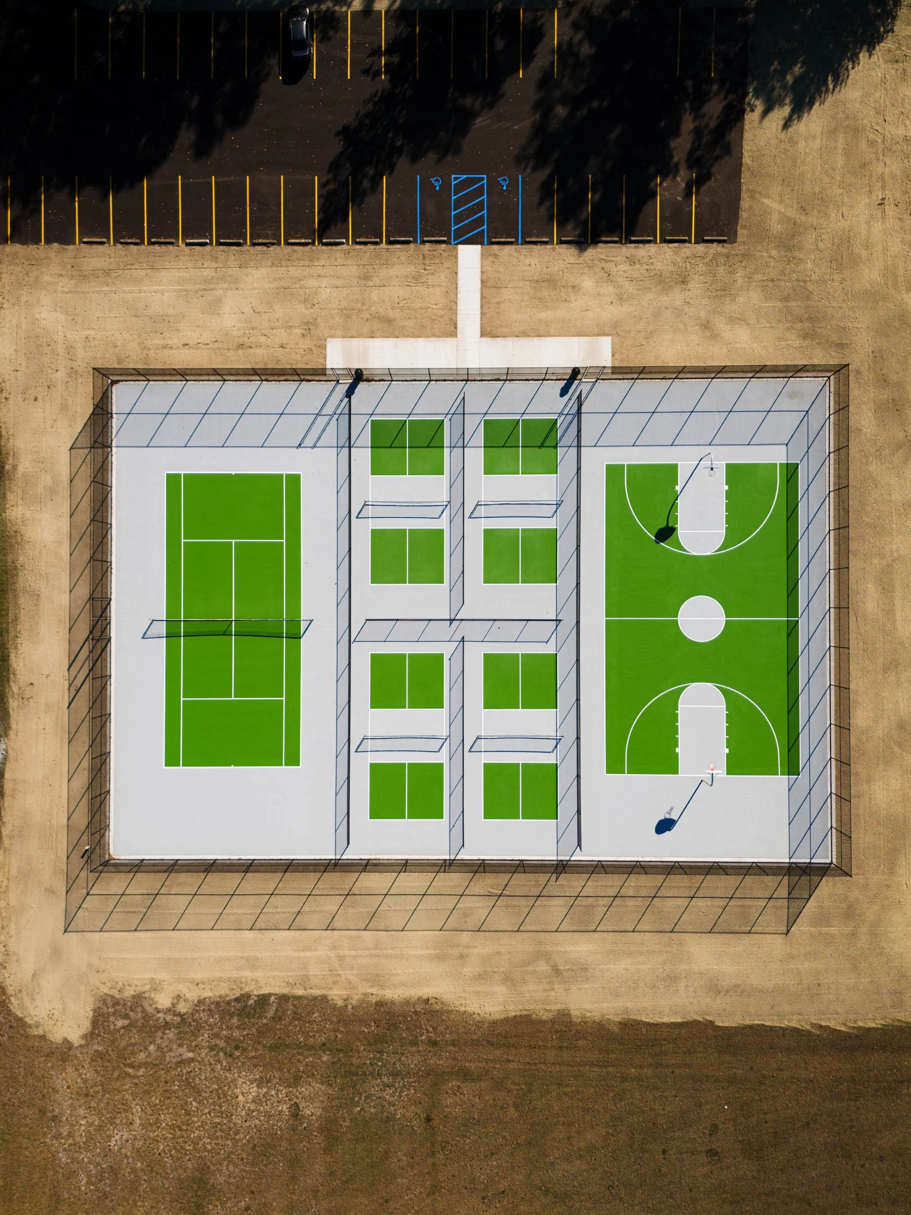Aerial view of a multi sports court in West Michigan with pickleball, tennis, and basketball courts, enclosed by a fence, with parking and trees surrounding the area.