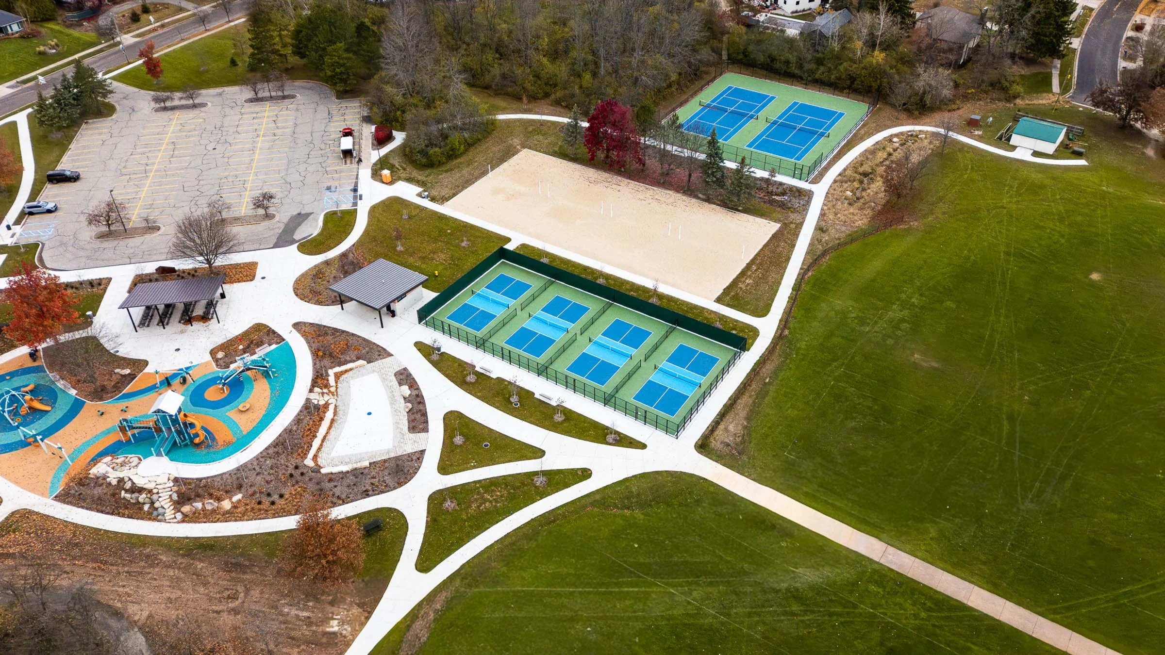 An aerial view of a park with playground equipment, tennis courts, and open grassy fields. The playground area has colorful surfacing and play structures, with walking paths surrounding it. There are two covered seating areas near the playground, and