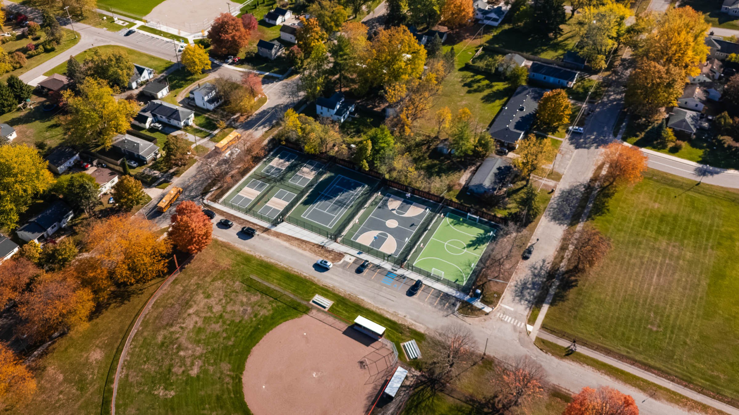 An aerial view of a neighborhood park showing multiple sports courts, including tennis courts, basketball courts, and a soccer field, surrounded by trees with colorful autumn foliage.