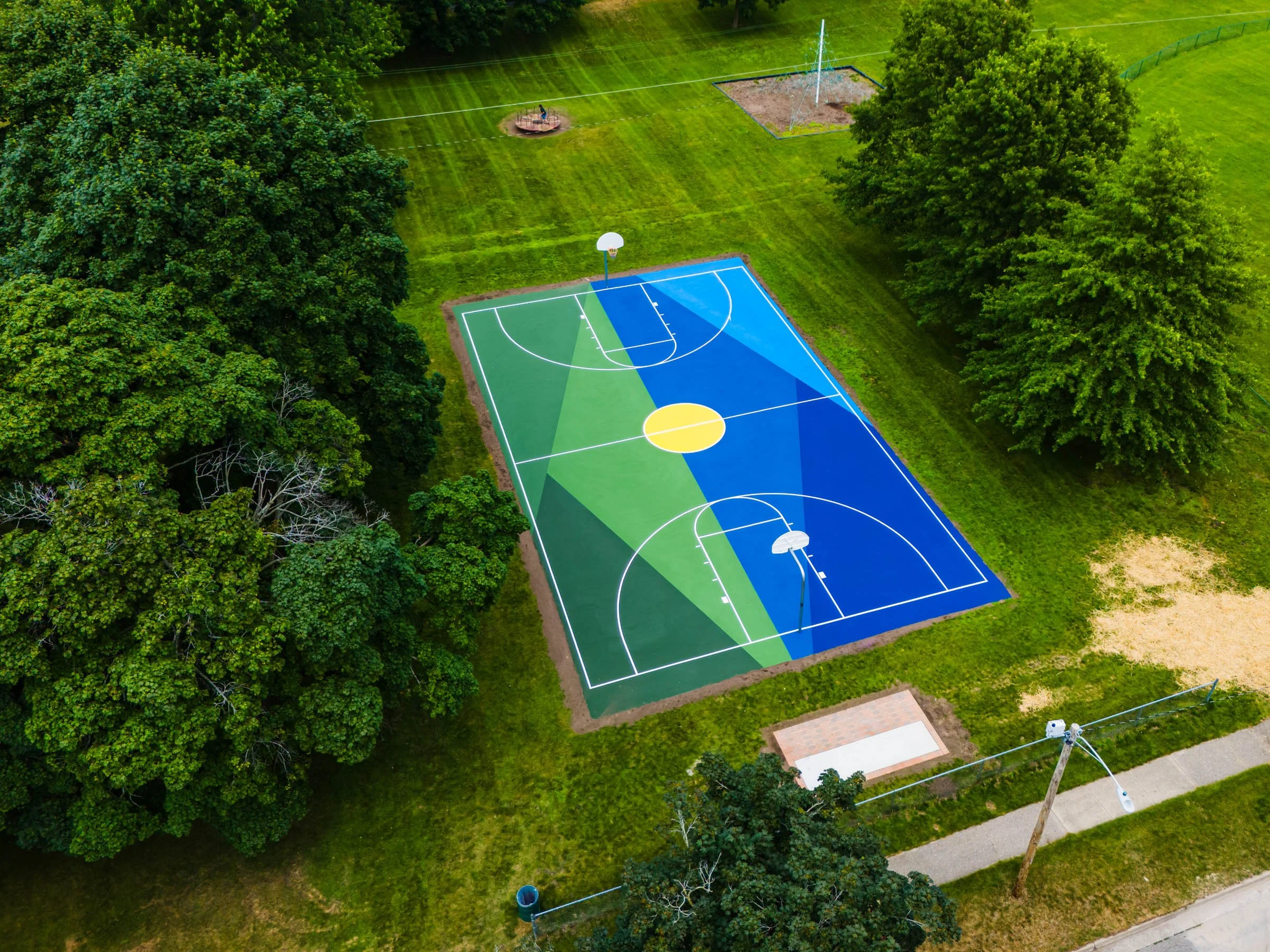 Colorful outdoor basketball court, in Grand Haven Michigan, with green, blue, and yellow painted sections, surrounded by green trees and grassy areas, with a white basketball hoop and a small paved area nearby.