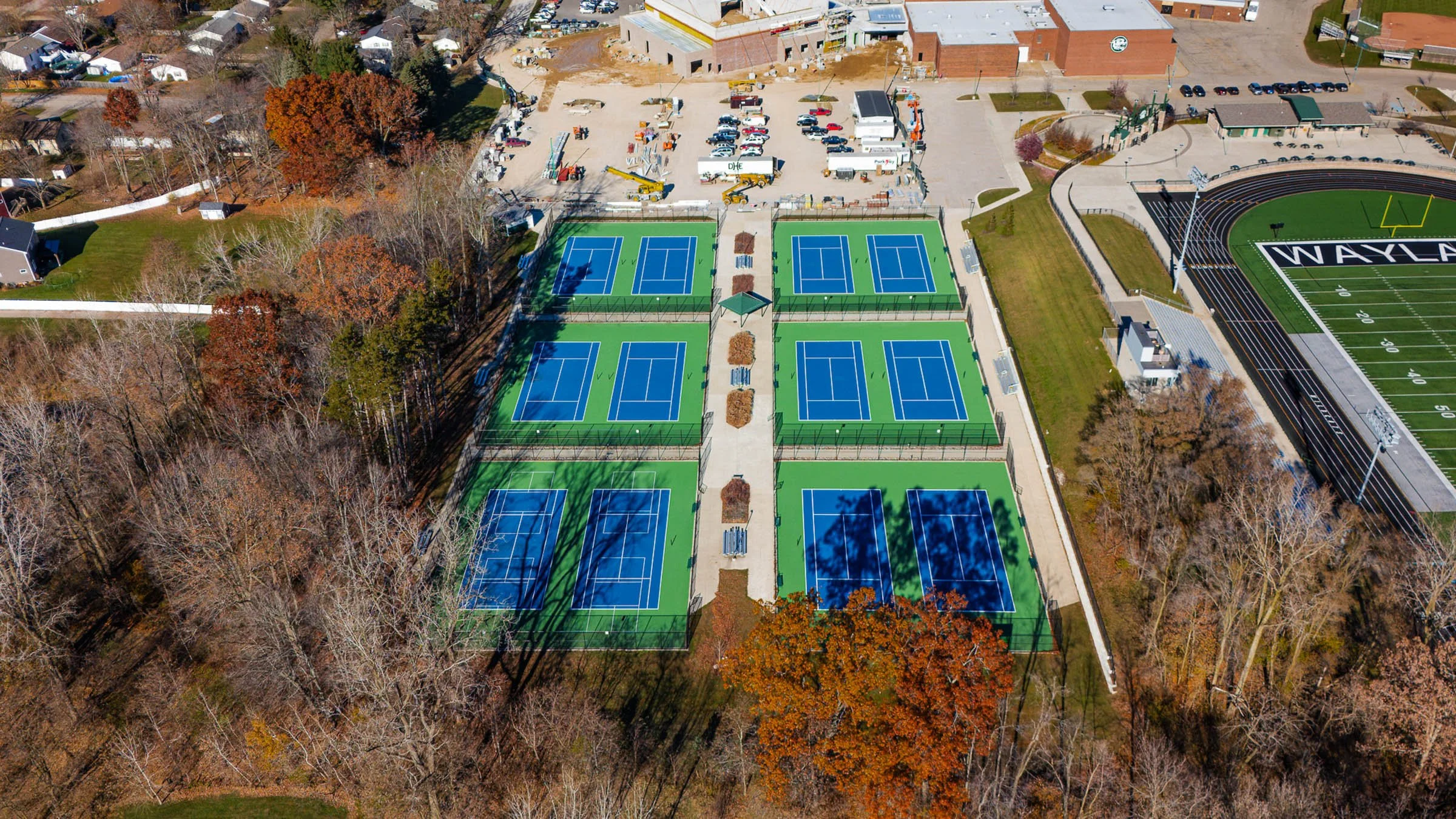 Aerial view of a sports complex with twelve blue and green tennis courts, a football field, and surrounding trees with autumn foliage.