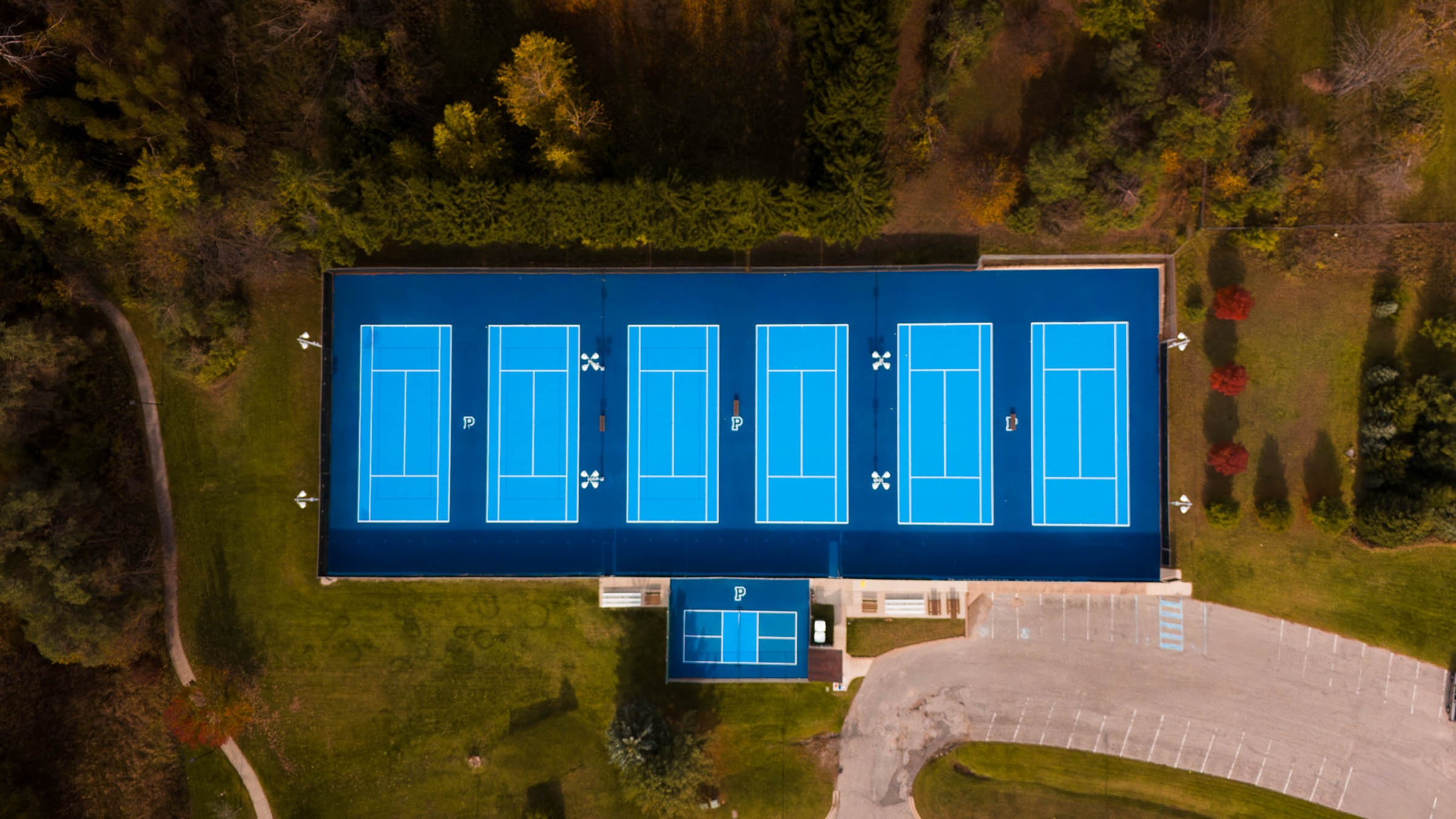 An aerial view of a blue outdoor tennis court facility in Petoskey Michigan featuring six courts, surrounded by greenery and parking lot.