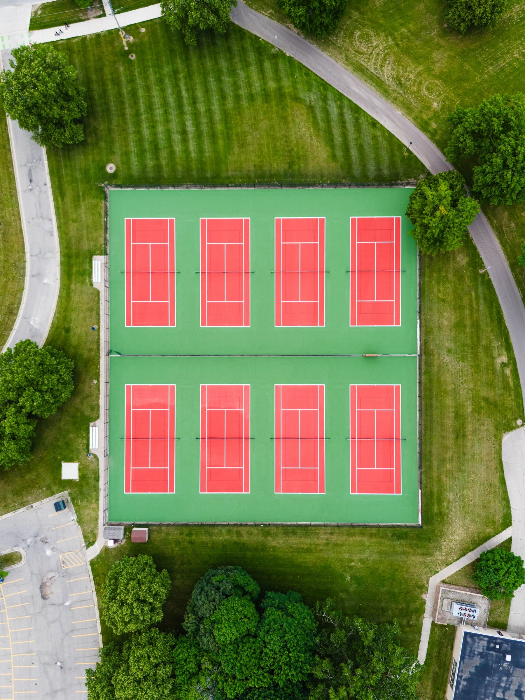 An aerial view of a tennis court complex with eight red courts separated into two sections, surrounded by green grass, trees, and walking paths.