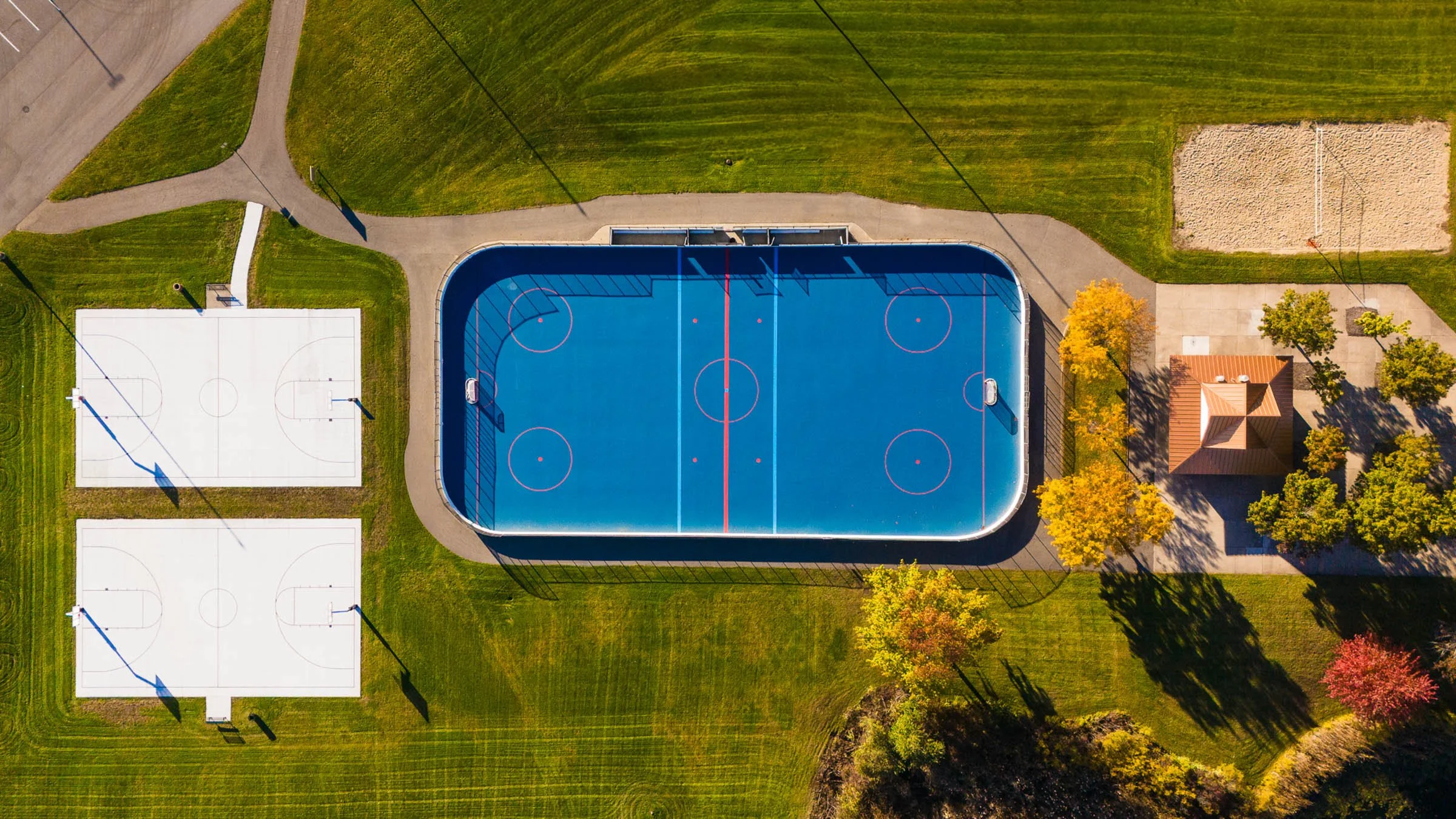 Aerial view of a sports complex, in Walker Michigan, with a large ice hockey rink, two basketball courts, a sand volleyball court, and a small building surrounded by trees in autumn.