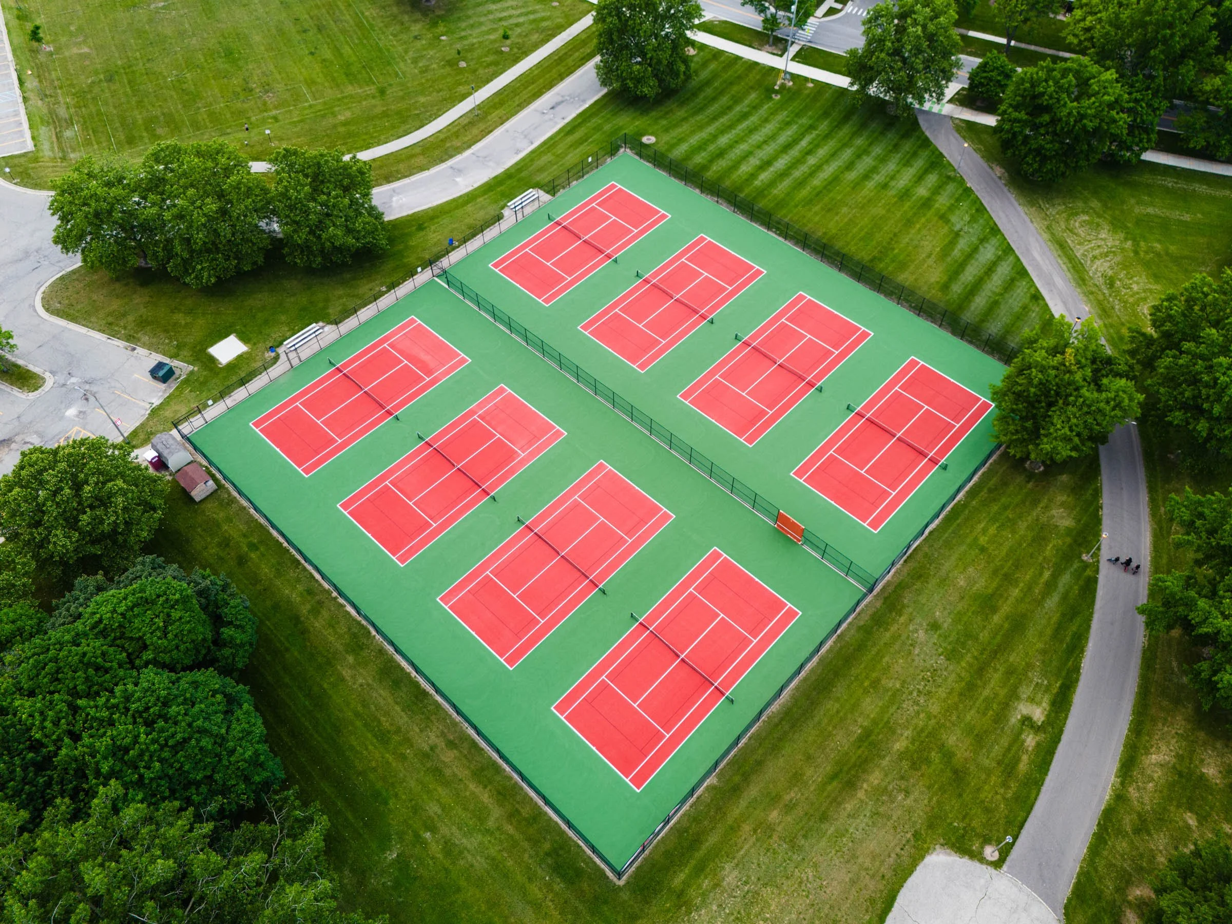 Aerial view of a tennis court complex with eight red courts surrounded by green fencing, situated within a park with trees, walking paths, and grassy areas.