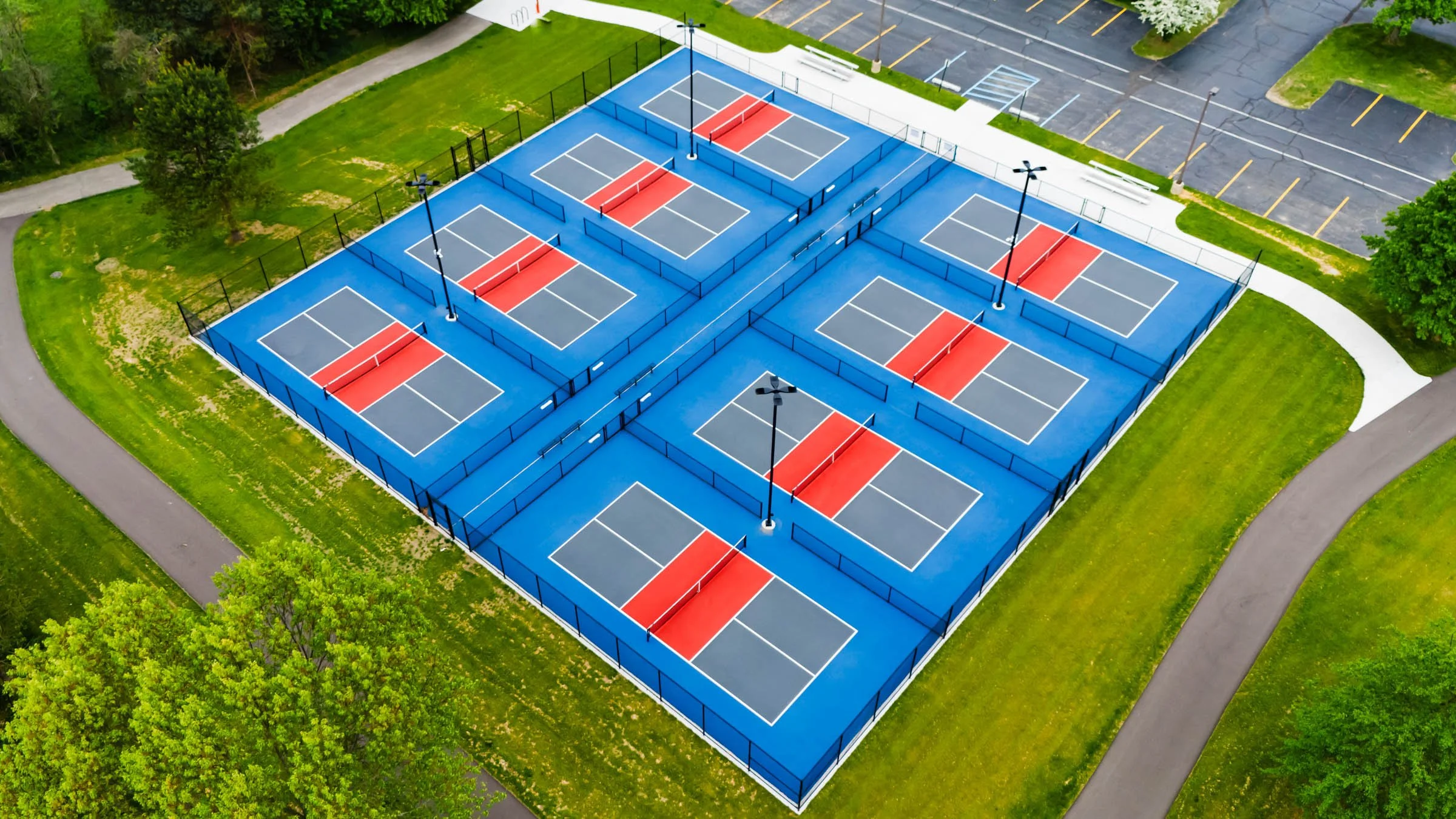 Aerial view of multiple blue and red tennis courts surrounded by green grass and trees, with a parking lot and pathways nearby.