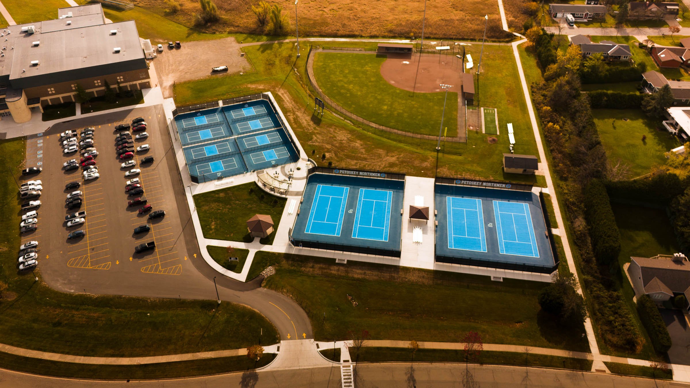 A school sports complex with multiple tennis courts and a baseball field. The tennis courts are painted blue with white lines, and the baseball field has a dirt infield and green grass outfield. There is a parking lot with several cars to the left of