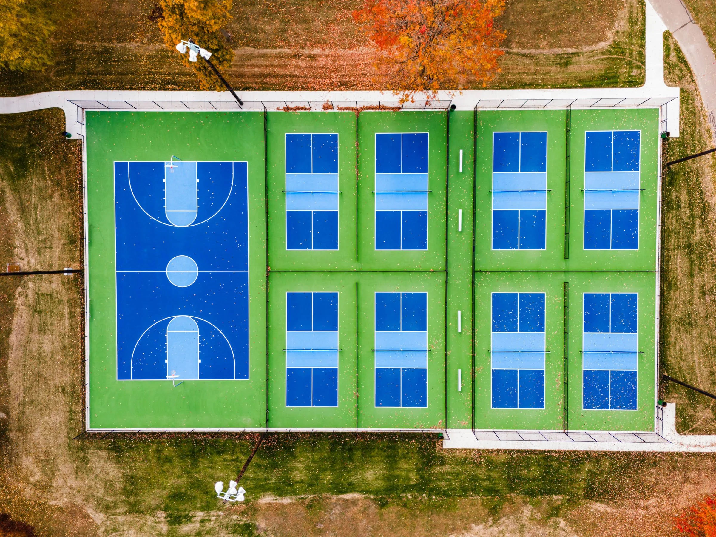 An aerial view of multiple outdoor basketball and pickleball courts. The courts have bright green surfaces with blue playing areas, white lines, and are surrounded by grass and pathways.