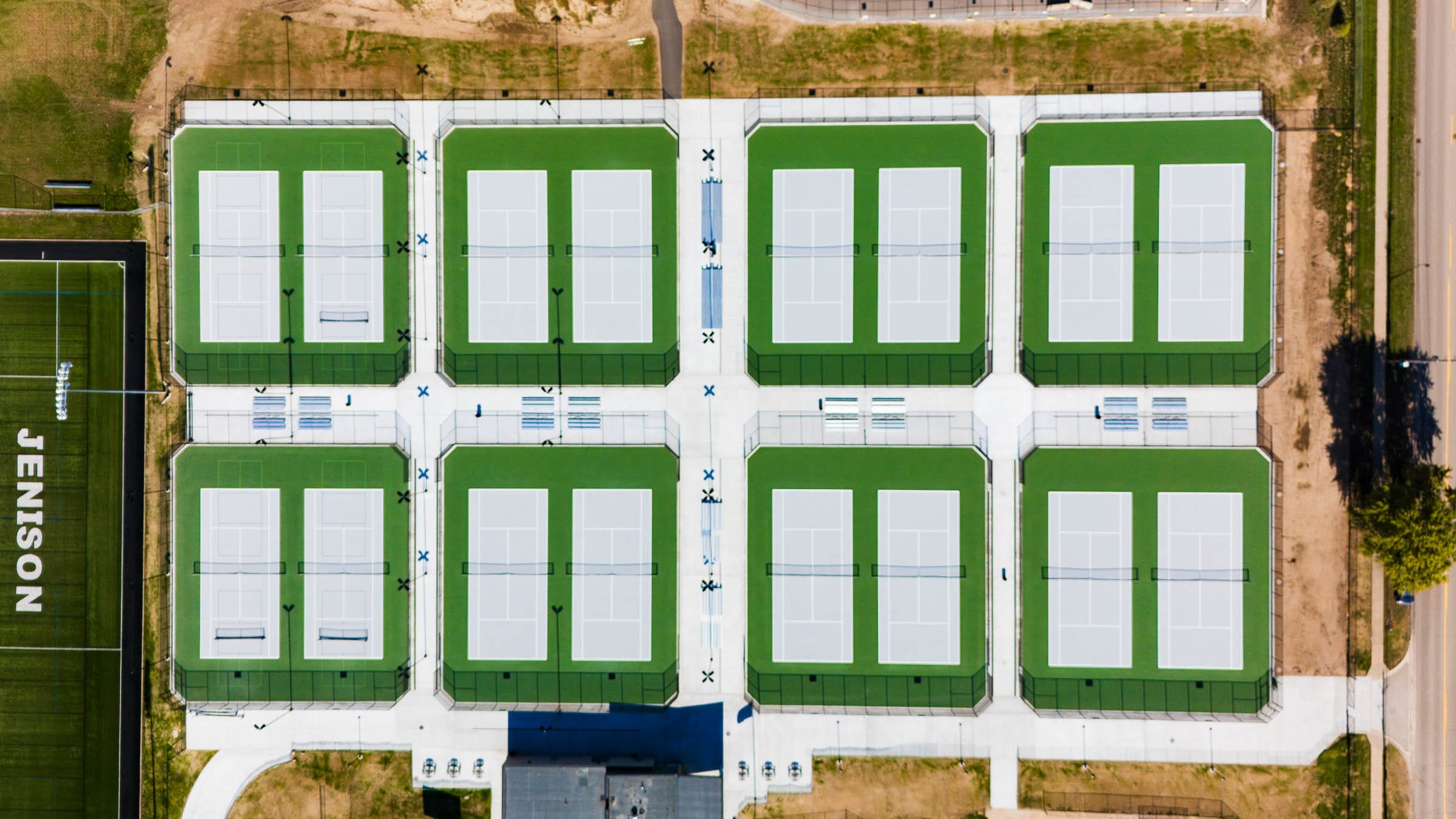 Aerial view of twelve outdoor tennis courts, in Jenison Michigan, with green surfaces and white lines, separated by concrete pathways, with a sandy area and trees nearby.