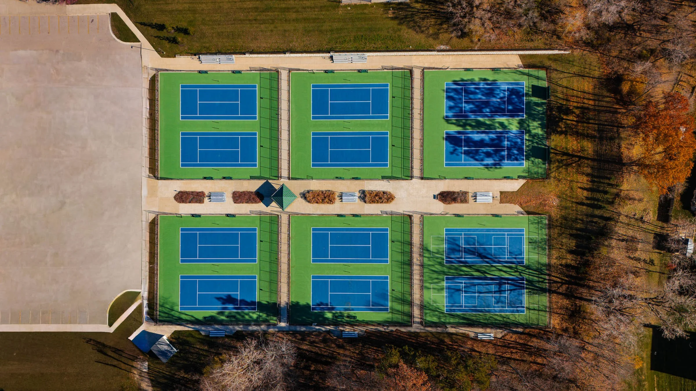 Twelve Tennis courts, in Holland Michigan, enclosed by chain-link fences. The courts are separated by walkways with benches and landscaped areas, surrounded by trees with autumn-colored leaves.