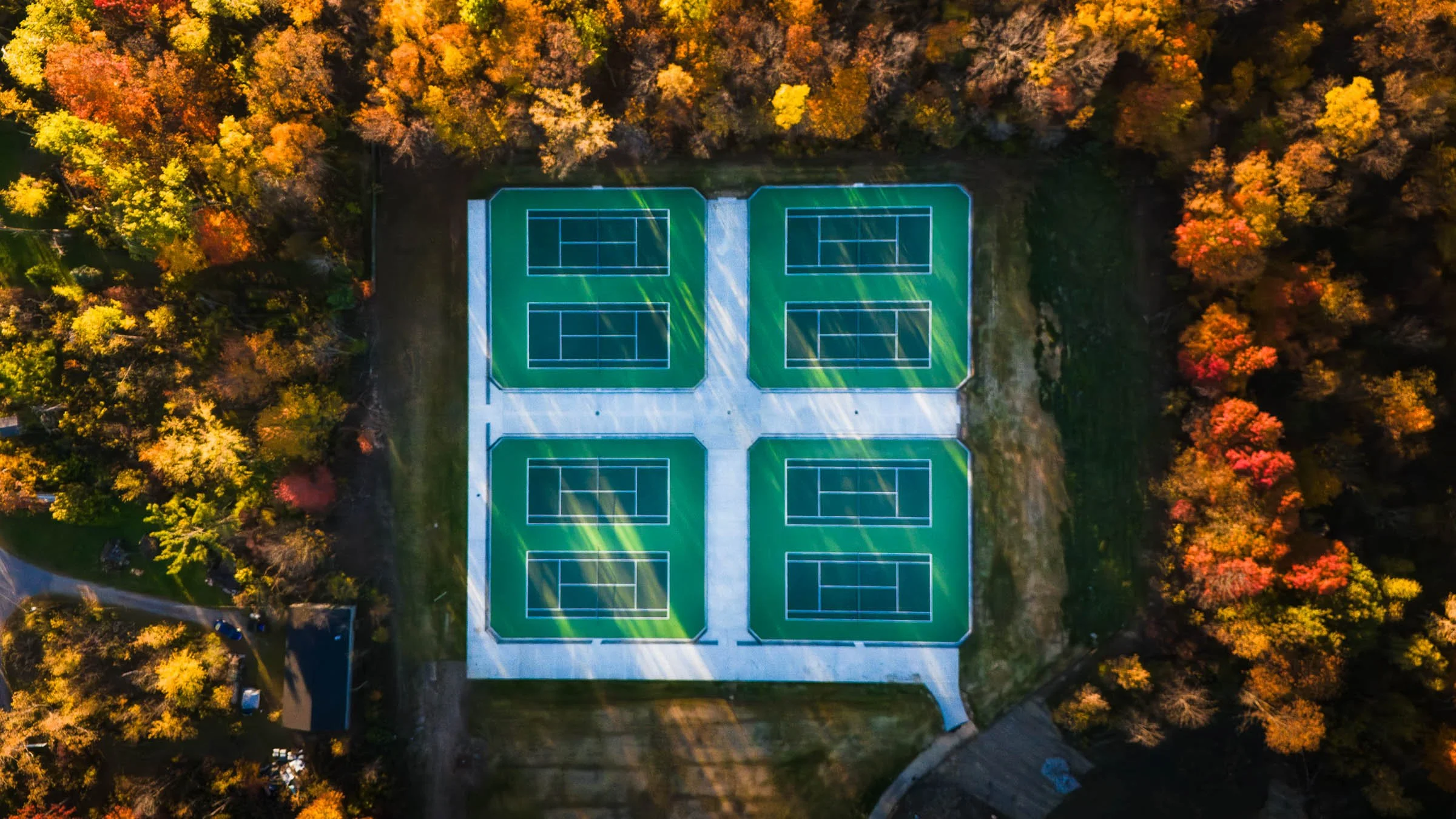 Comstock Park High School post tension concrete tennis courts with two green playing surfaces and white lines, surrounded by trees with autumn foliage, aerial view.