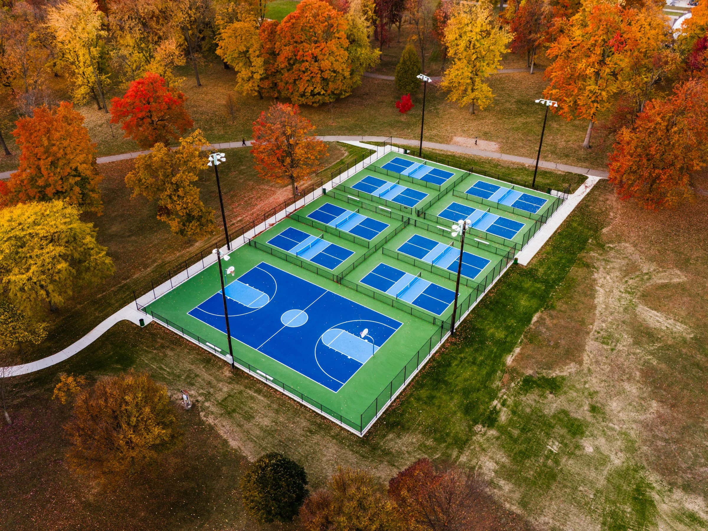 An aerial view of a pickleball court complex surrounded by trees with fall foliage, featuring multiple blue pickleball courts, some separated by green partitions, with tall lights around the courts and a walking path nearby.