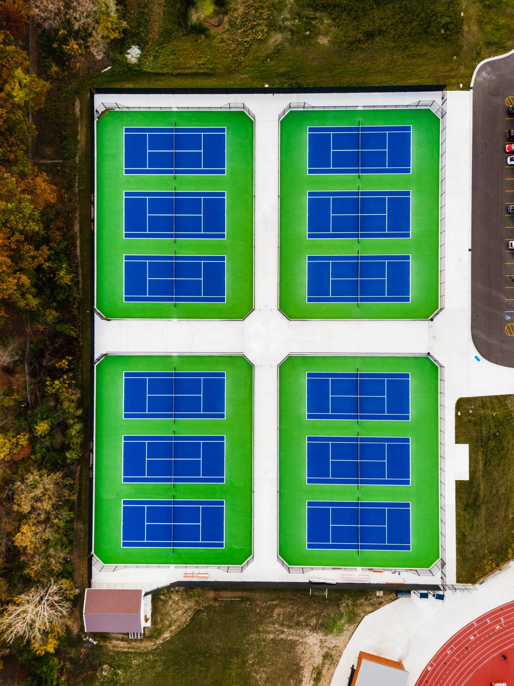 Aerial view of twelve blue and green tennis courts, in Rockford Michigan, with blue playing surfaces separated by concrete walkways, surrounded by trees and a parking lot.